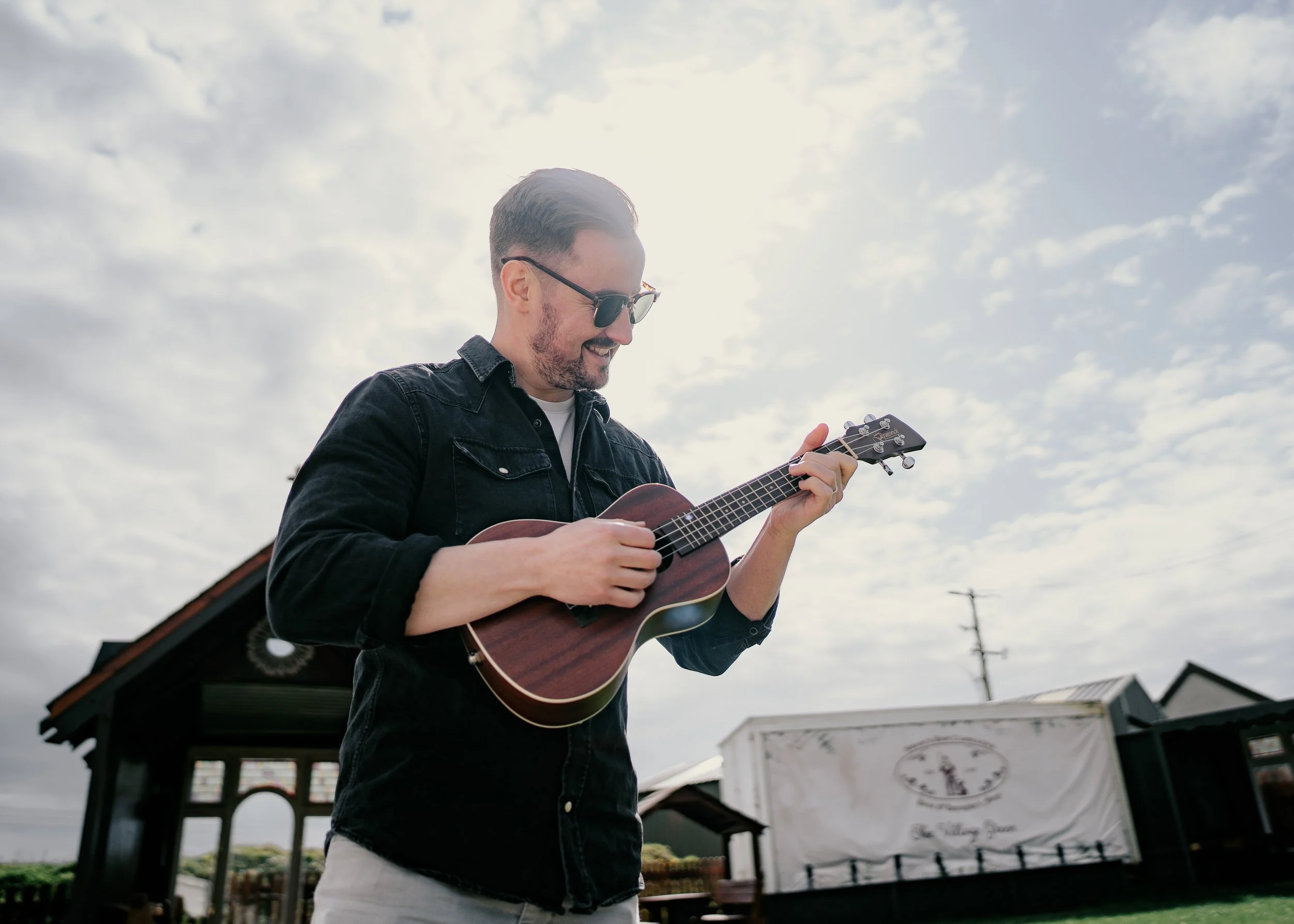 A man wearing sunglasses and a black denim jacket is smiling and playing a ukulele outdoors under a cloudy sky.