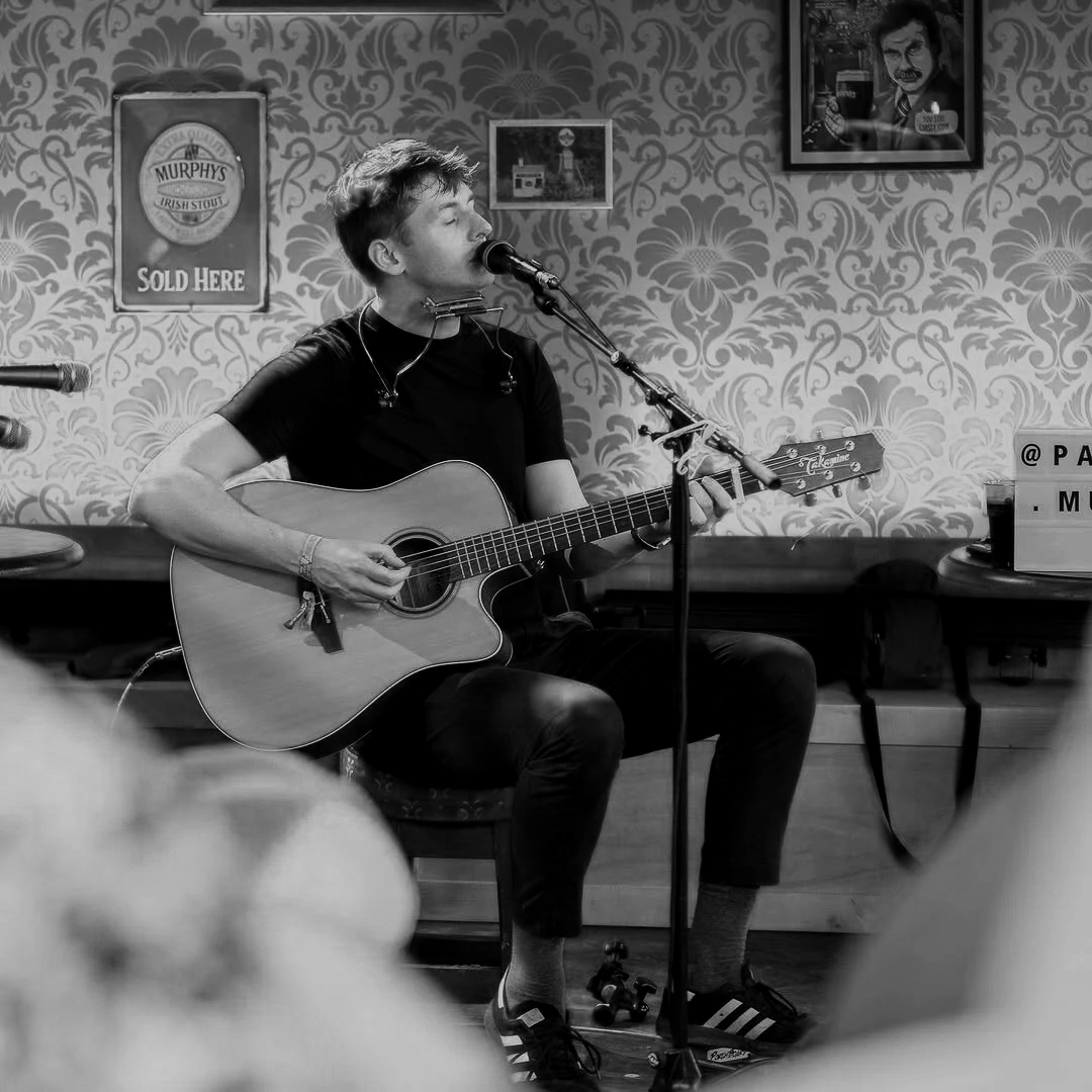 A young man playing an acoustic guitar and singing into a microphone on a small stage in a cozy pub or bar. The background features patterned wallpaper, framed pictures, and a Guinness sign. The scene is in black and white.