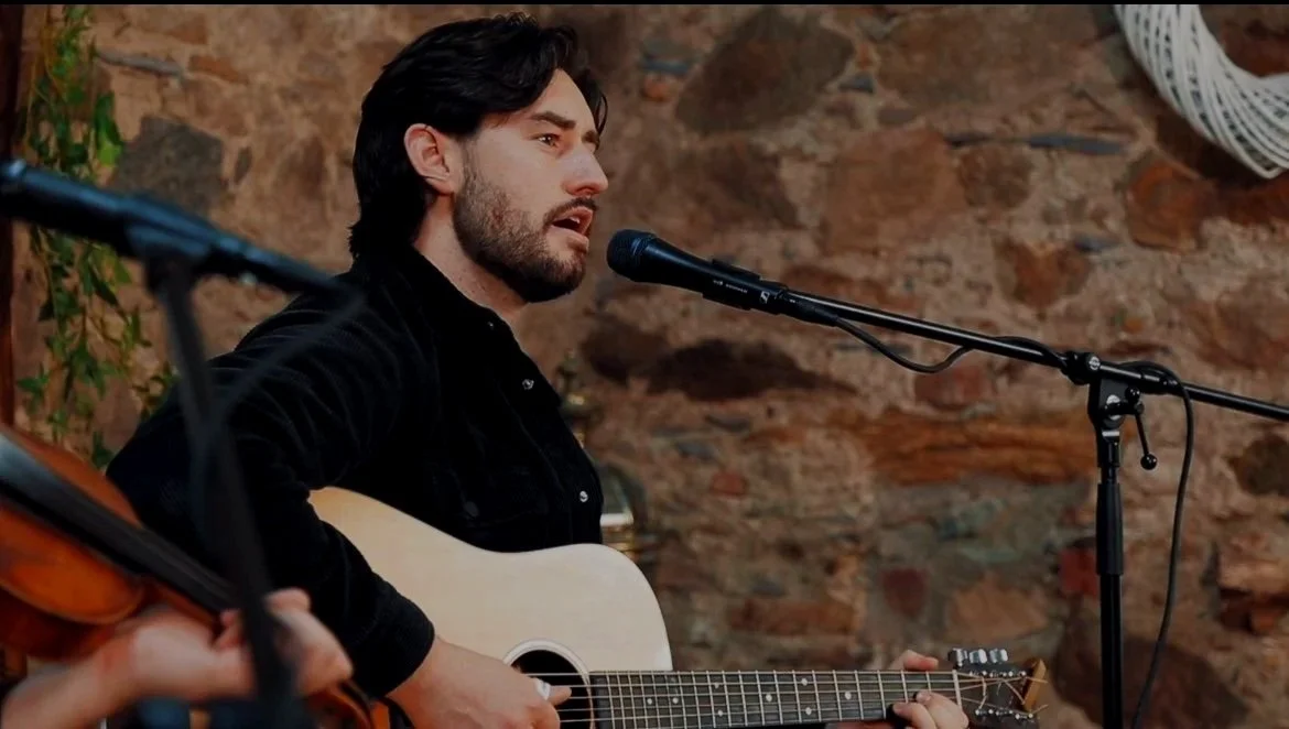 A man with dark hair and beard playing an acoustic guitar and singing into a microphone, sitting in front of a brick wall.