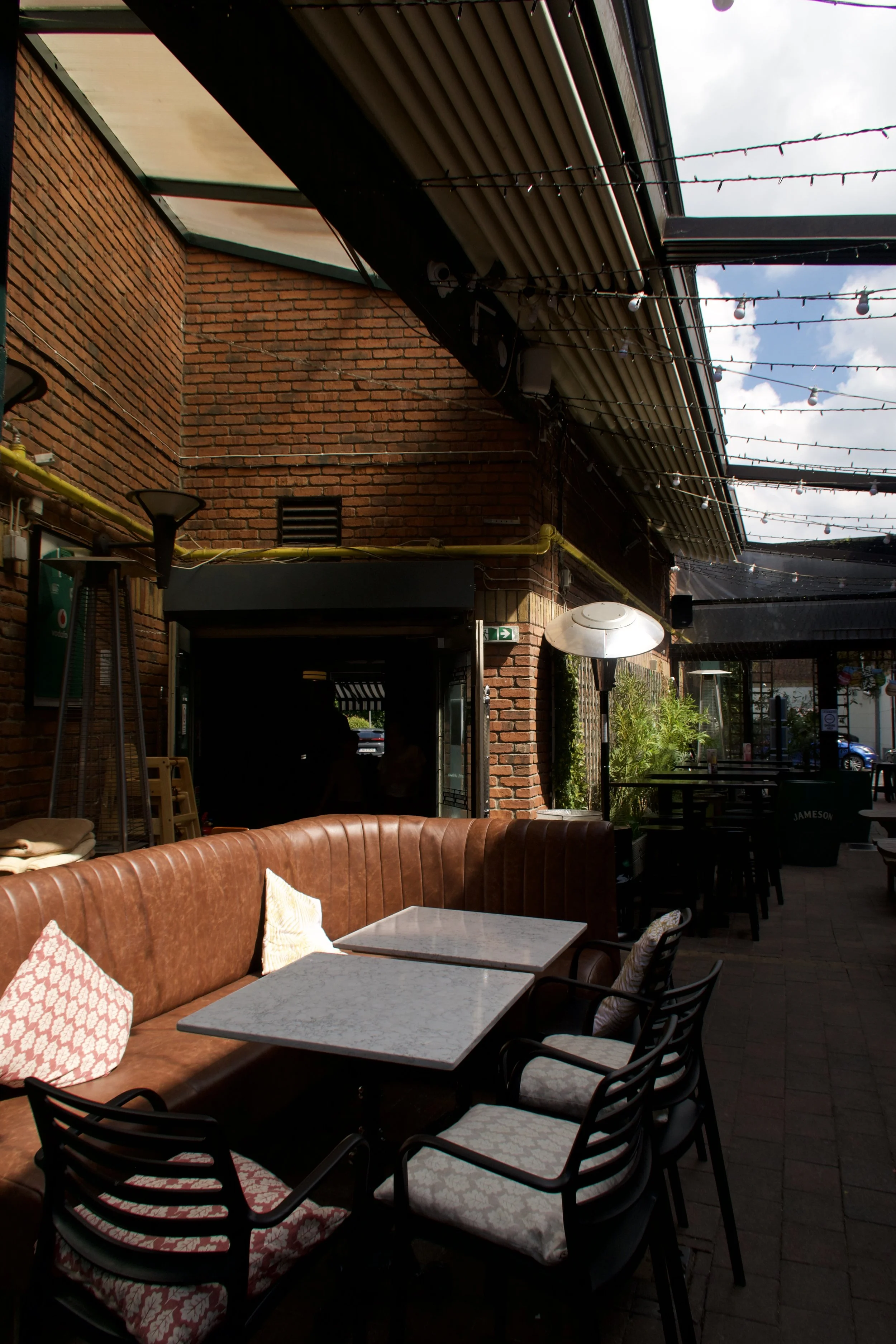 Outdoor patio area with brick walls, string lights overhead, and a mix of seating including a leather sofa with patterned cushions, and black chairs around tables. Part of the sky with clouds is visible through the open roof structure.