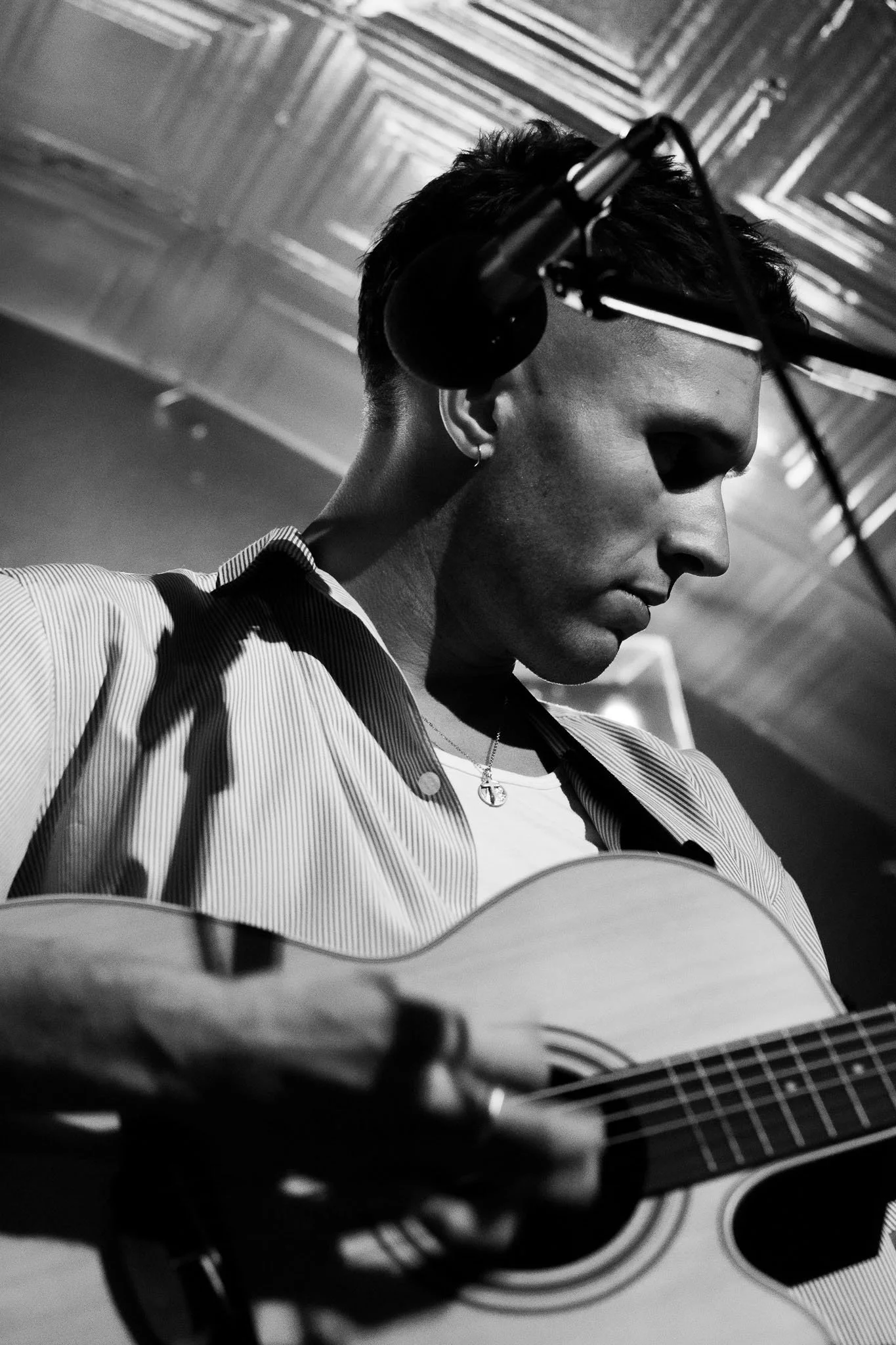 A young person with short hair playing an acoustic guitar in a recording studio, with headphones on and a patterned shirt, captured in black and white.