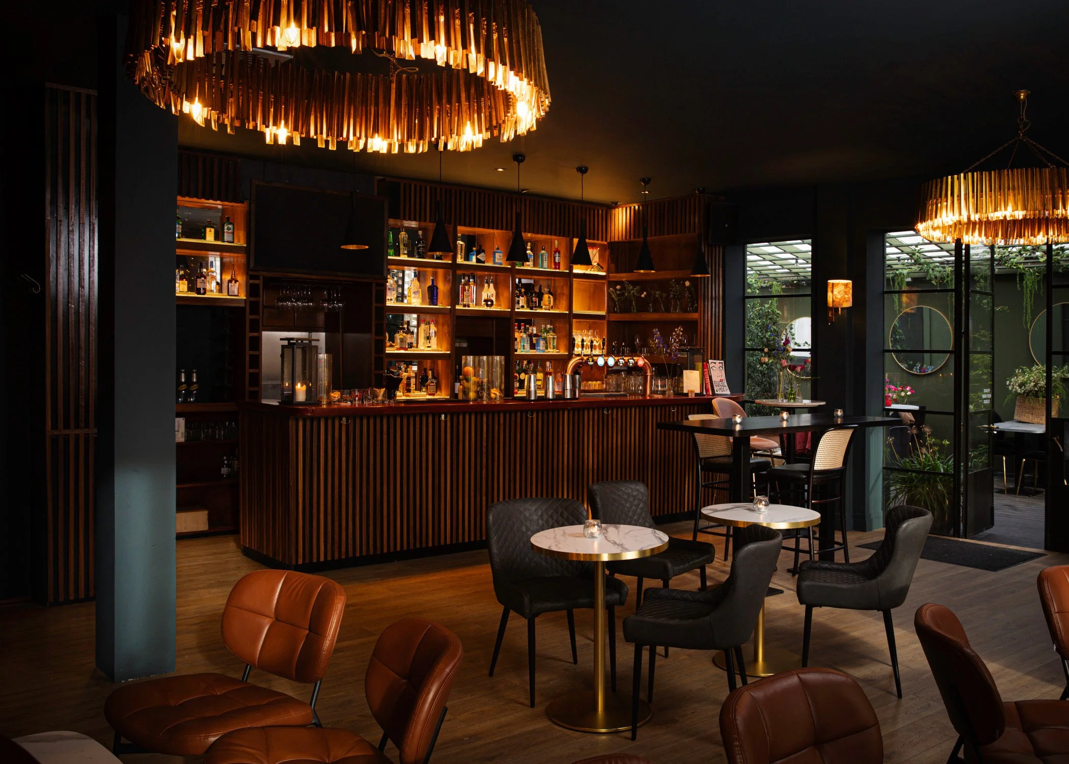 An outdoor patio area of a restaurant or café with brick walls, string lights, and various seating options including a leather bench with decorative pillows, tables, and black chairs. There is a partially open glass roof and a cloudy sky visible above.