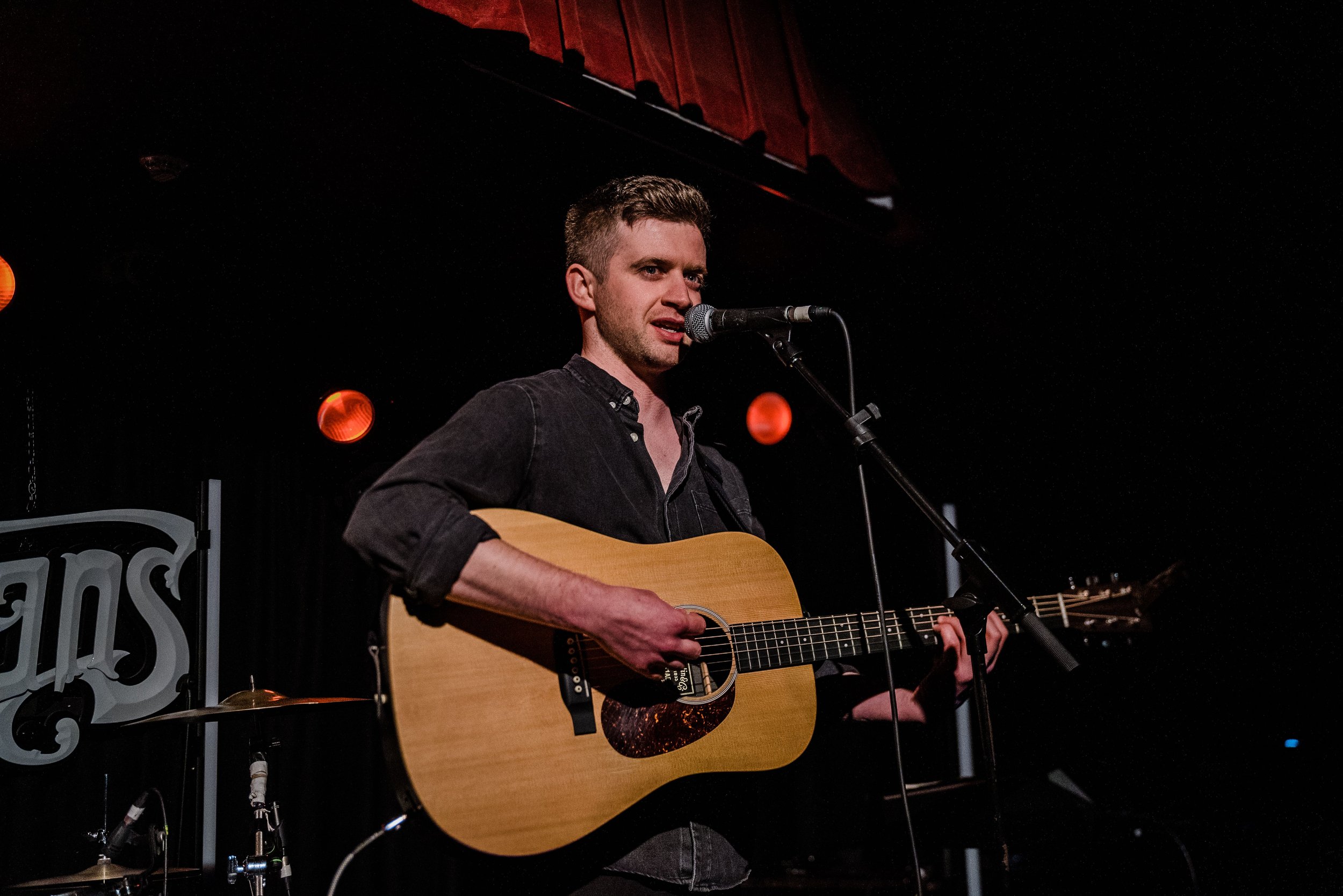 A man is performing on stage with a guitar and singing into a microphone in a dark venue with red stage lights.