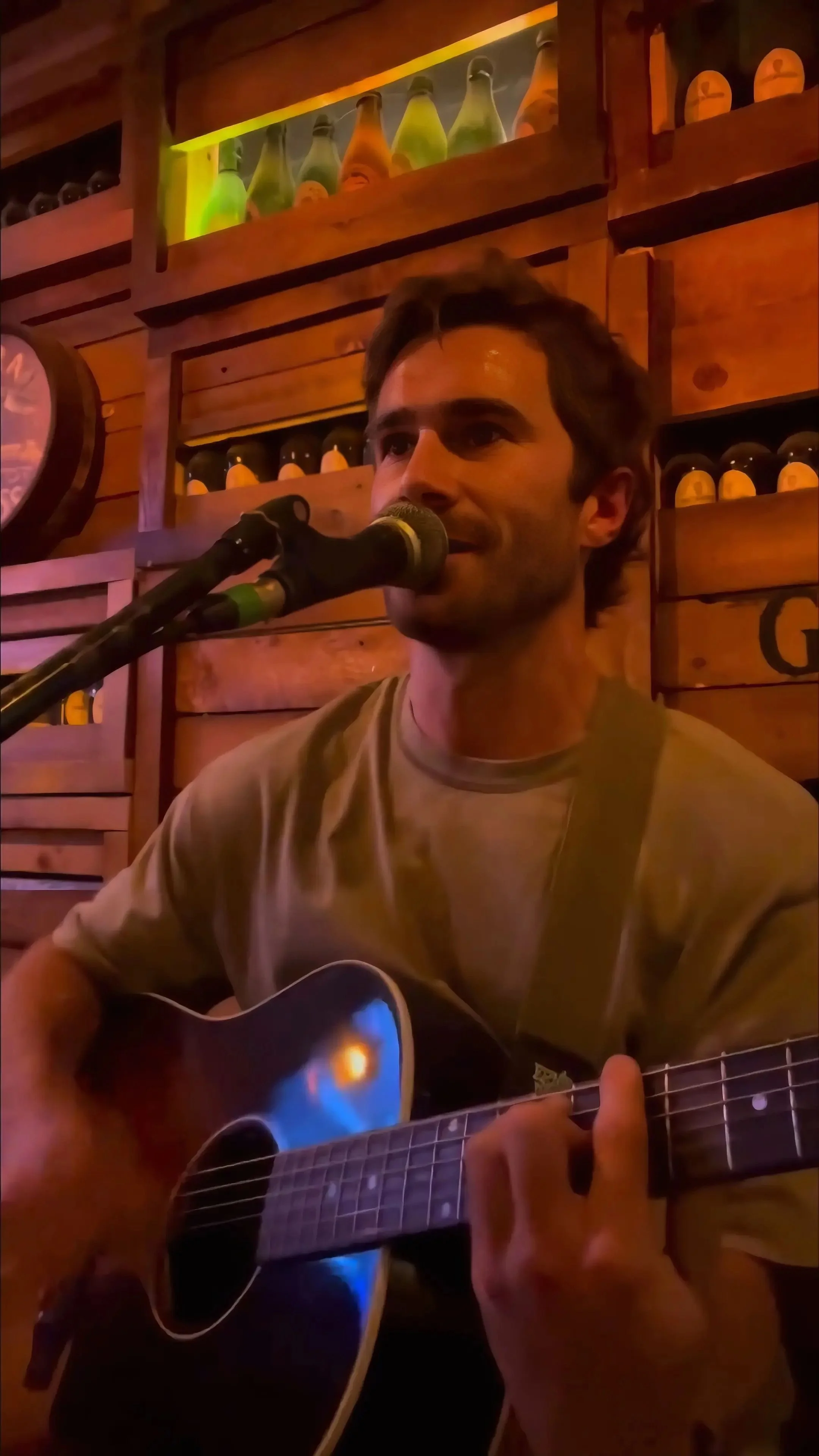 A man playing an acoustic guitar and singing into a microphone in a cozy, wooden-themed venue.