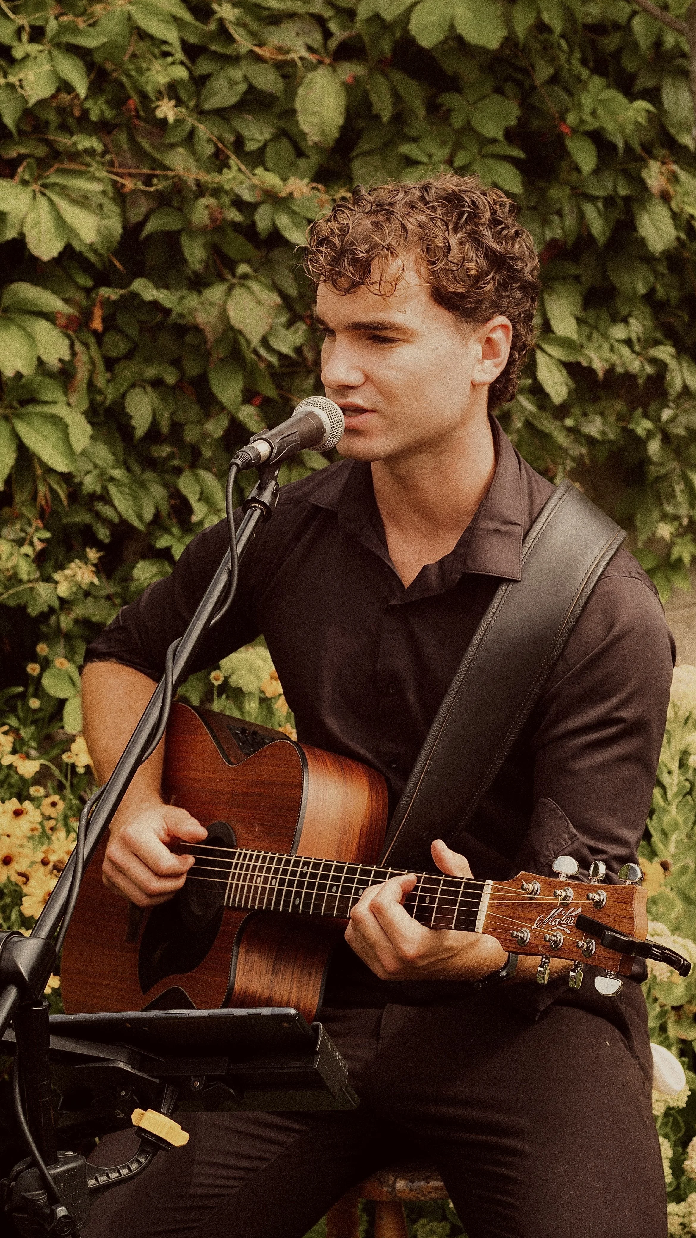 A young man with curly hair wearing a black shirt, sitting outdoors with green foliage in the background, playing an acoustic guitar and singing into a microphone.