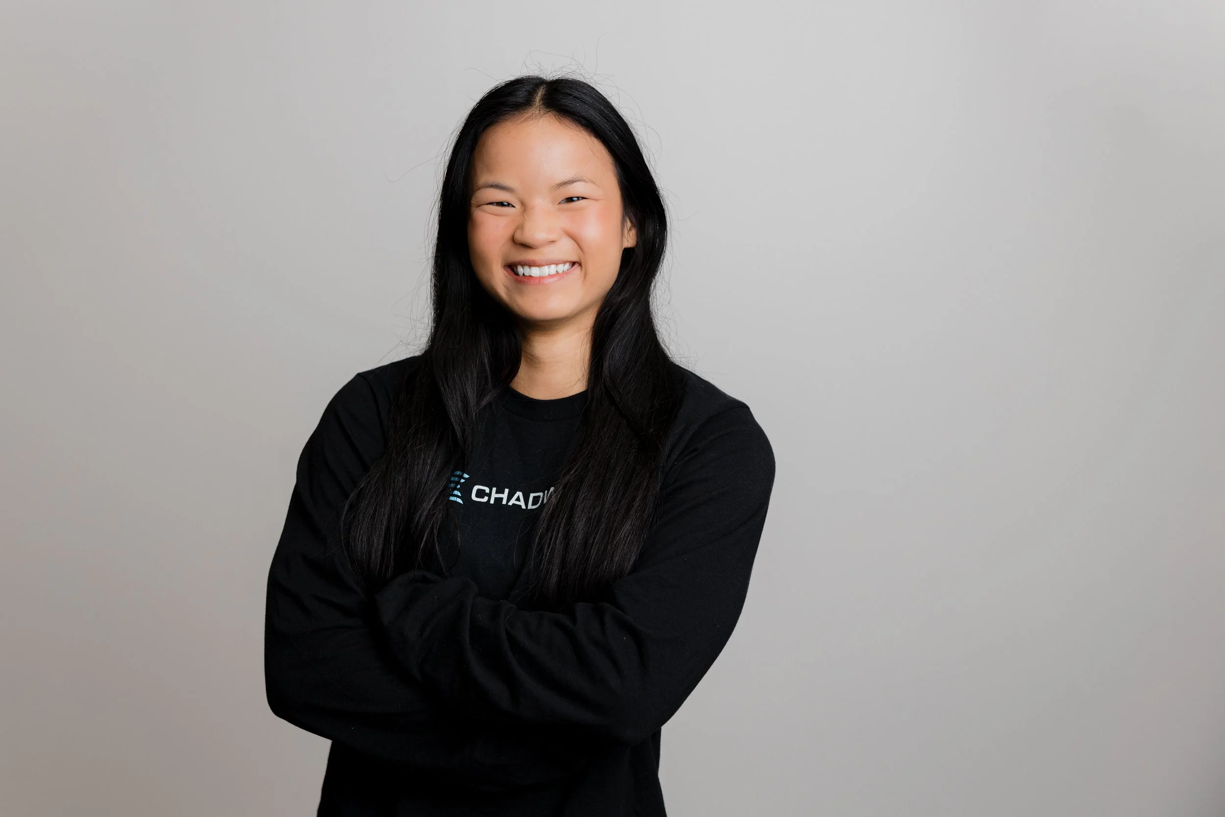 Young woman with long black hair smiling in a black long-sleeve shirt against a plain gray background.