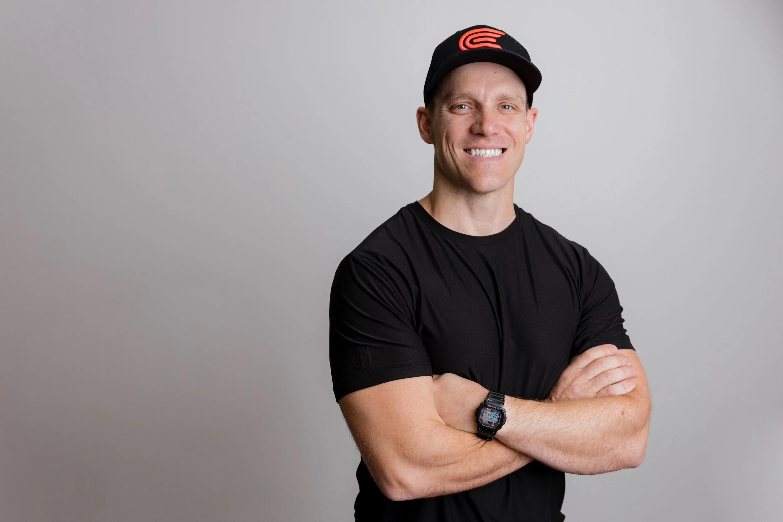 Smiling man in black t-shirt and cap with crossed arms against a plain background.