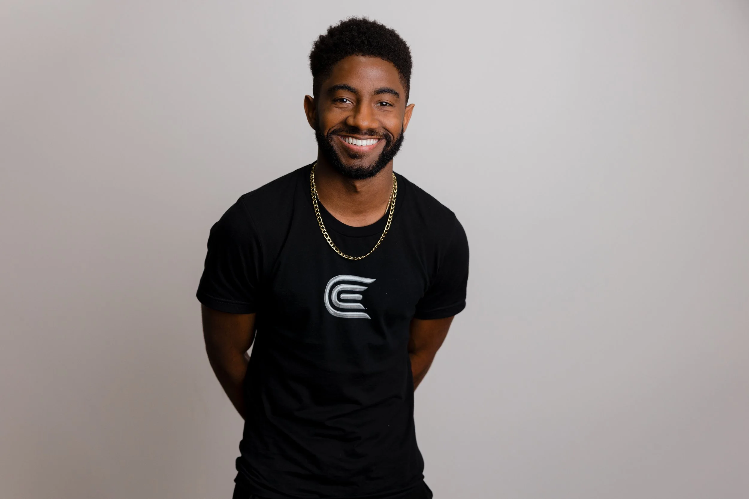 A smiling man with short curly hair, beard, and gold chain necklace, wearing a black t-shirt with a white logo, standing against a plain light gray background.