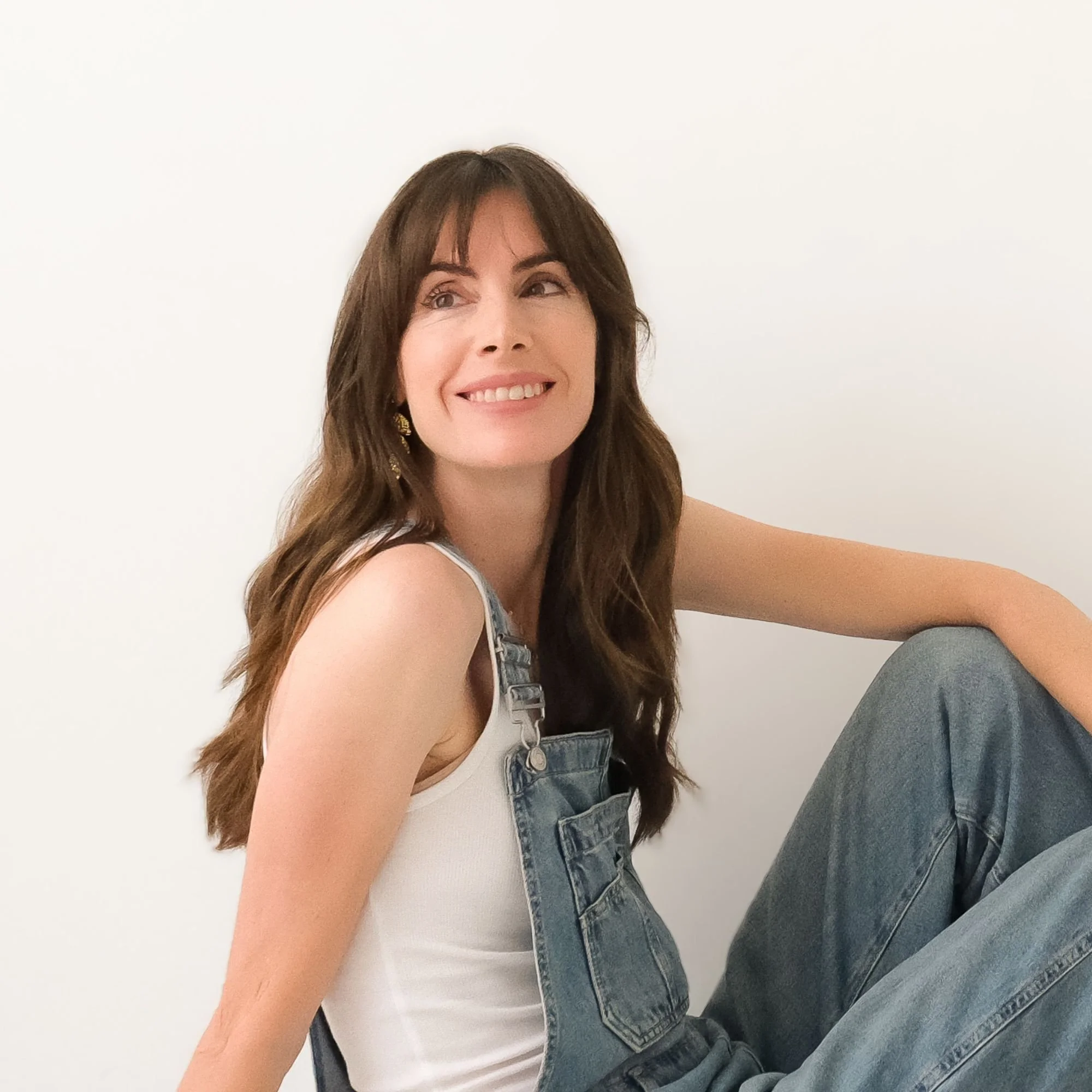 A young woman with long brown hair and light skin is smiling, sitting against a plain white wall. She is wearing a white tank top and denim overalls, and appears relaxed and happy.