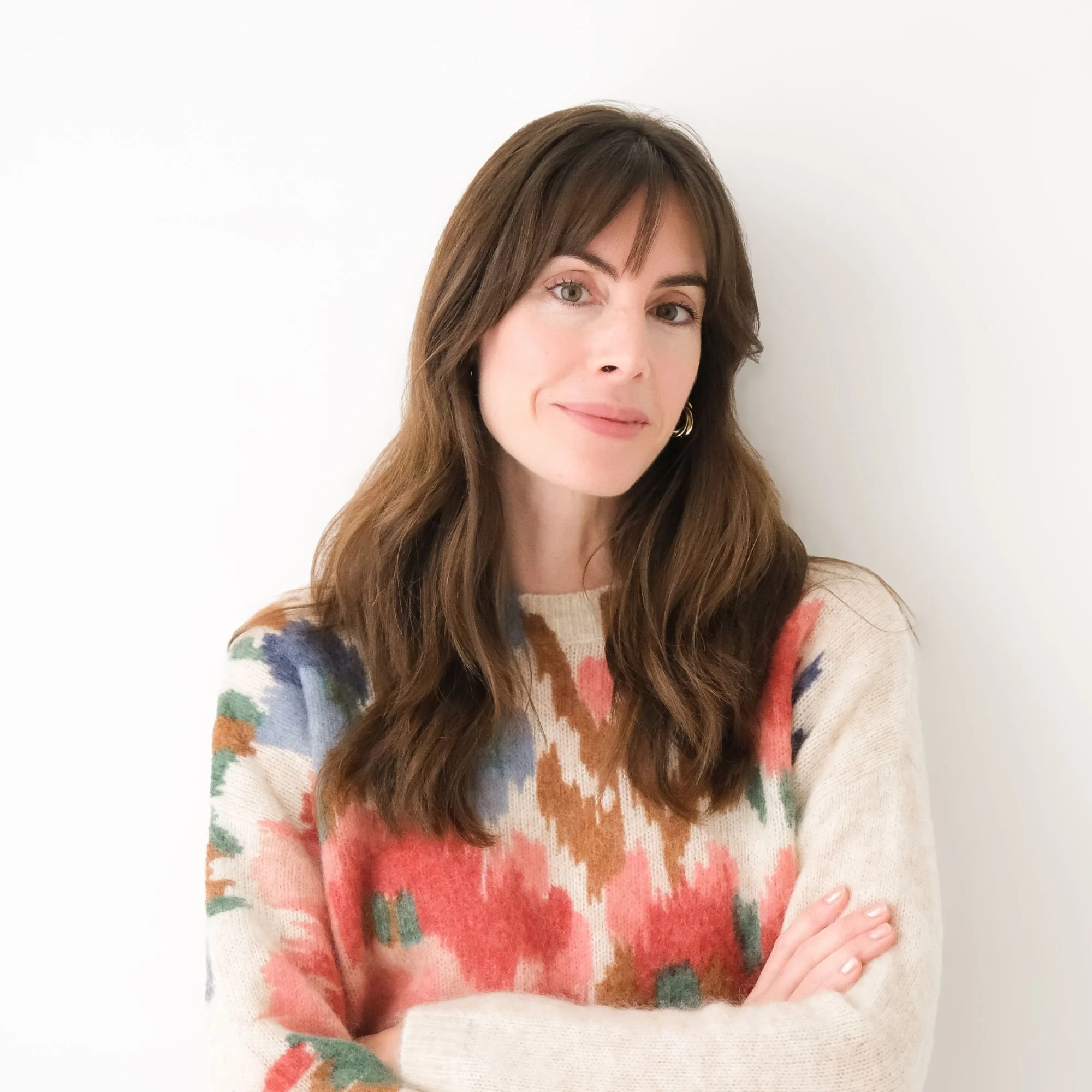 A woman with long brown hair wearing a colorful patterned sweater standing against a plain white wall. Keri-Lee Larmand.