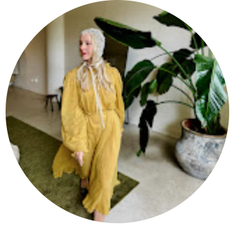 A woman with long gray hair wearing a yellow outfit, standing indoors near a potted plant.