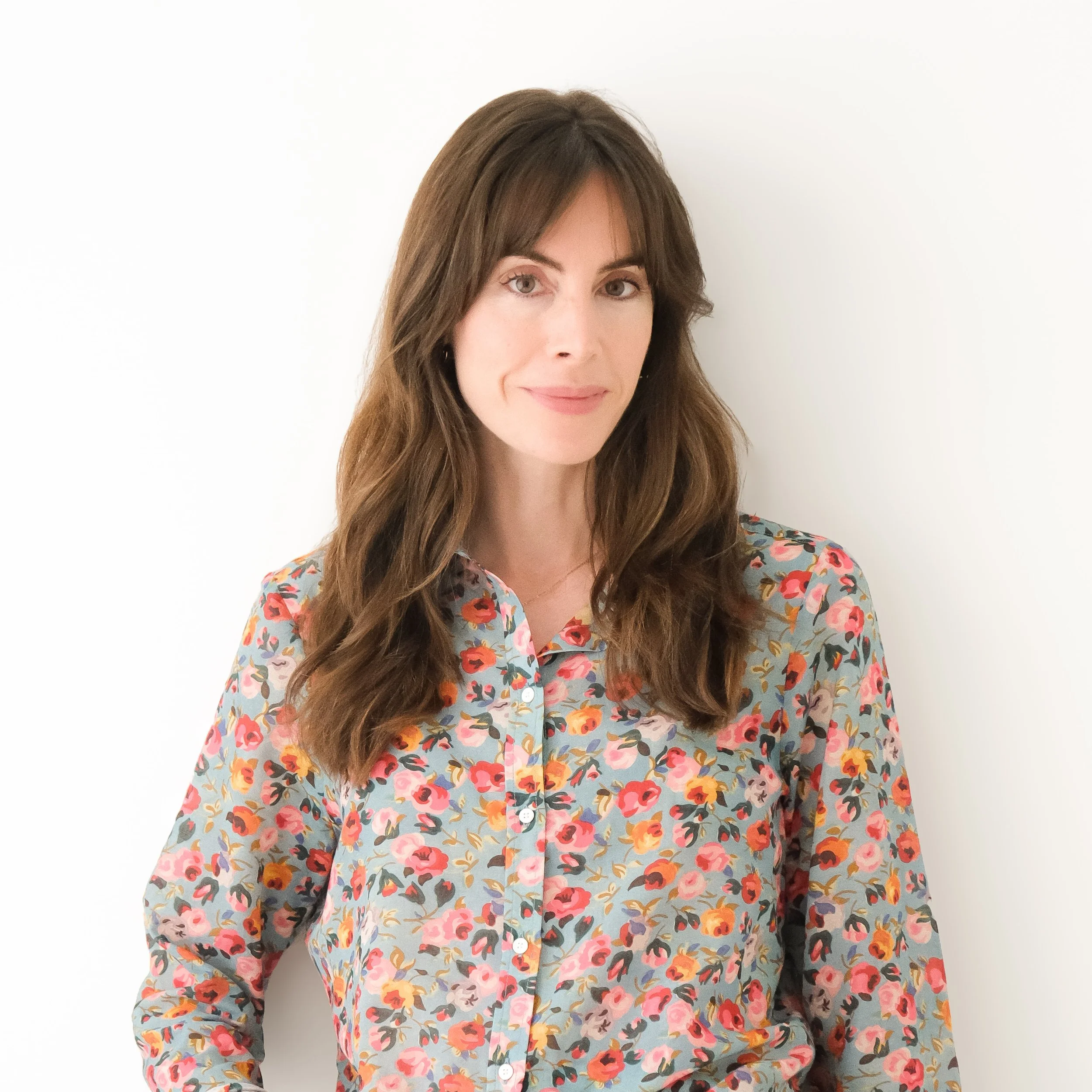 Woman with long brown hair wearing a floral blouse, standing against a plain white wall.