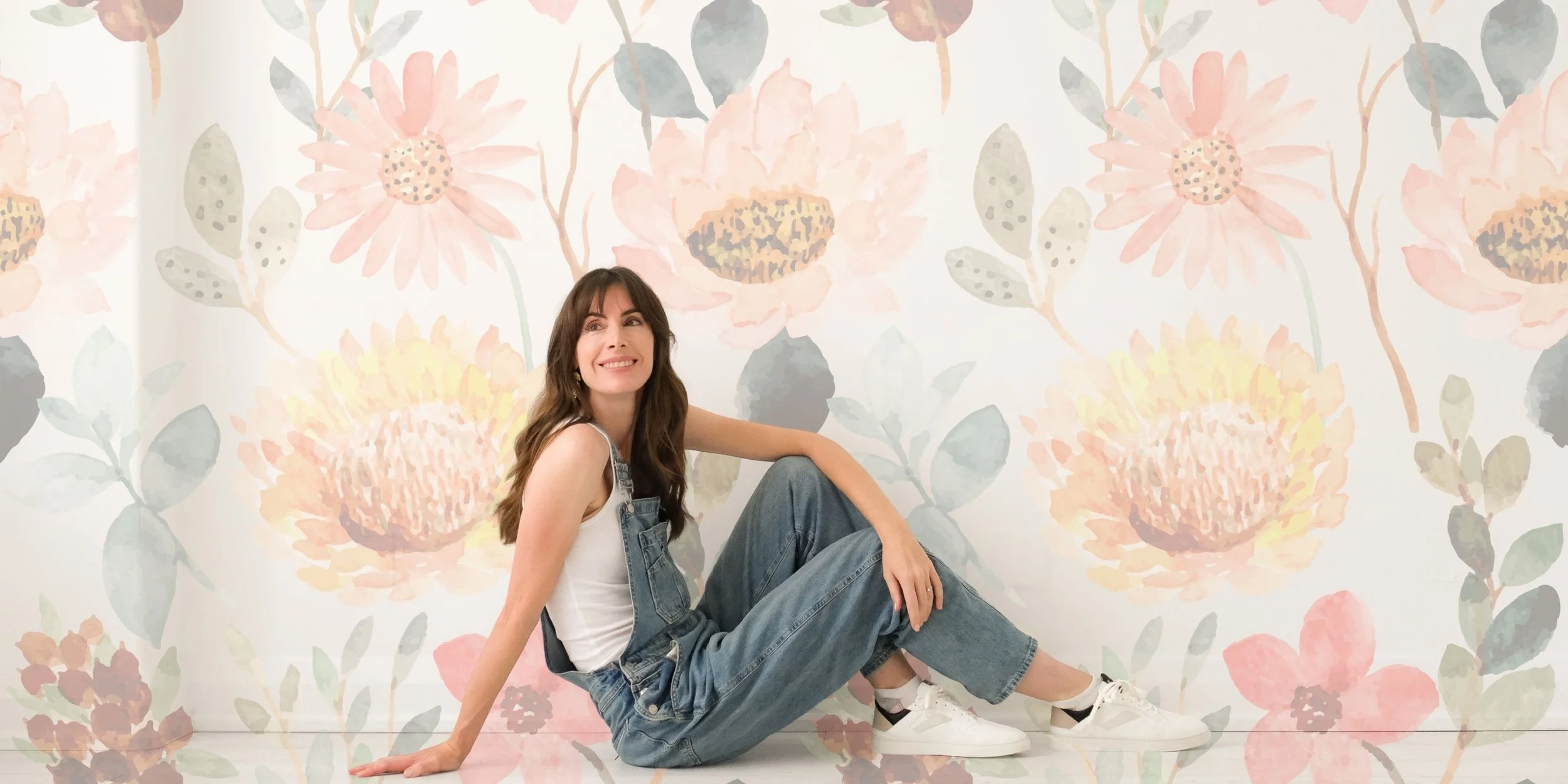 A woman with long brown hair sitting on the floor against a floral wall with pink, peach, and muted green watercolor flowers, wearing a white tank top, blue denim overalls, and white sneakers, smiling.