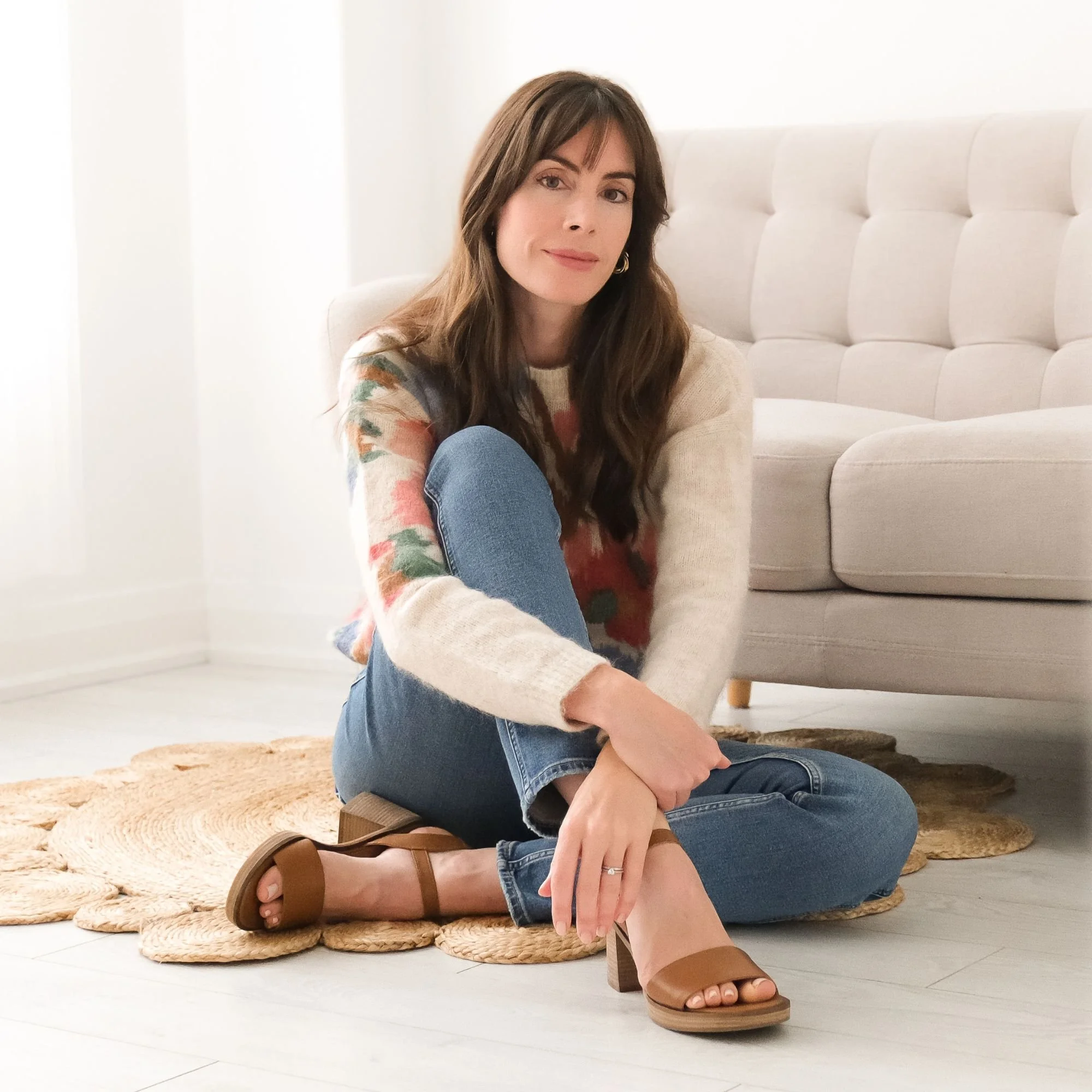 Woman sitting cross-legged on a woven rug, wearing a floral sweater, blue jeans, and tan heels, in a bright living room with a beige sofa in the background.