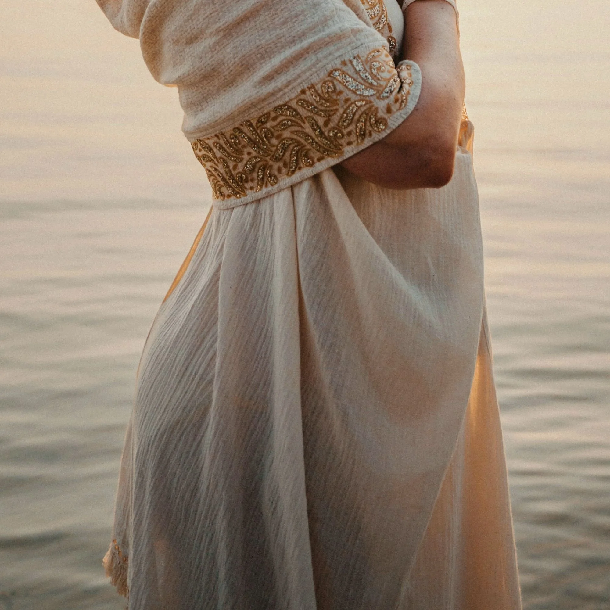A woman wearing a cream-colored dress with gold embroidery on the sleeve, standing by the water at sunset.