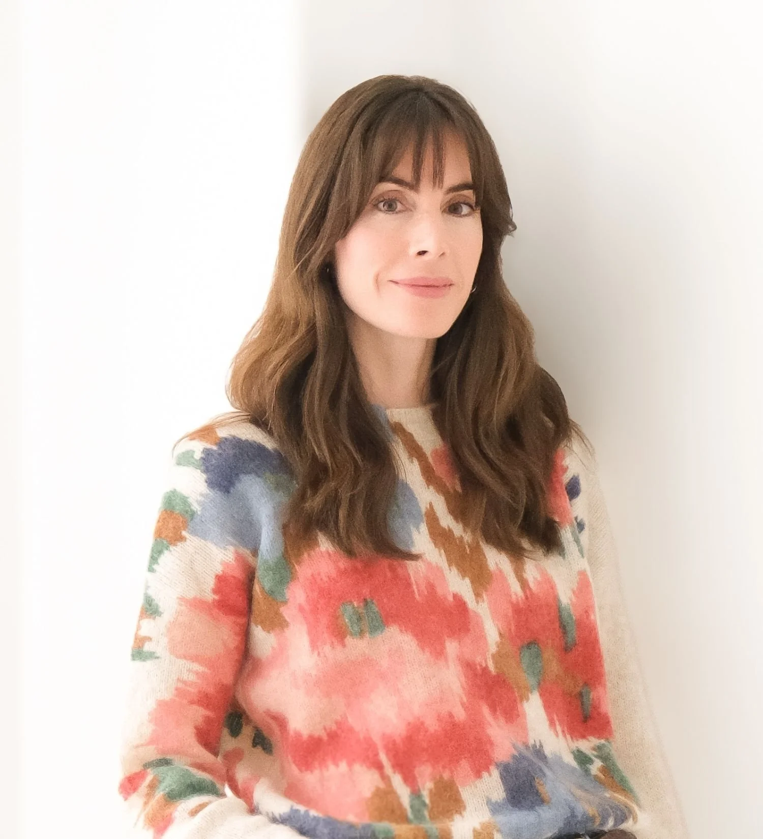 A woman with long, wavy brown hair, fair skin, wearing a multicolored abstract patterned sweater, standing against a plain white wall.