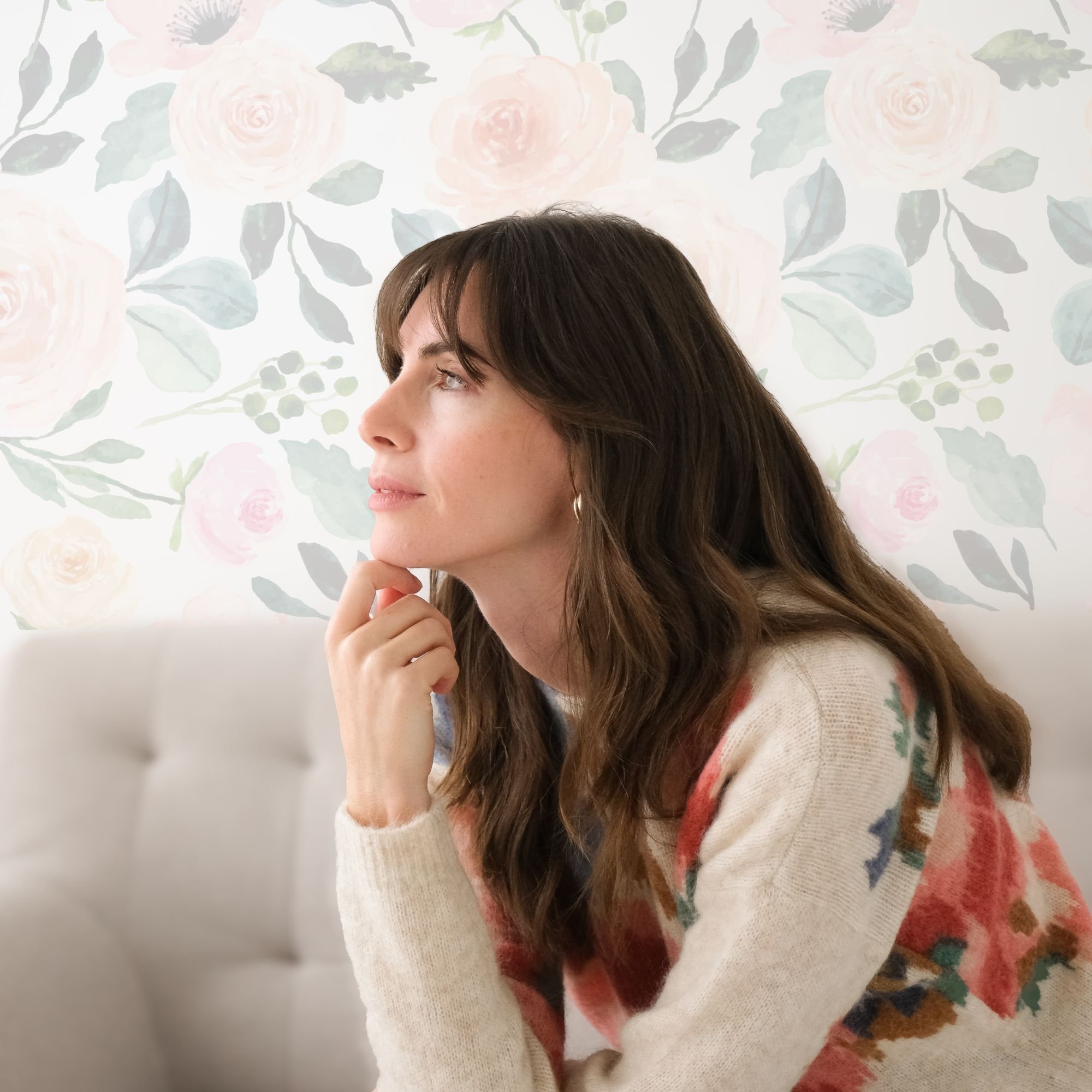 A woman with long brown hair, wearing a cream-colored sweater with floral patterns, sitting indoors with a floral wallpaper background, resting her chin on her hand and looking thoughtfully to the side.