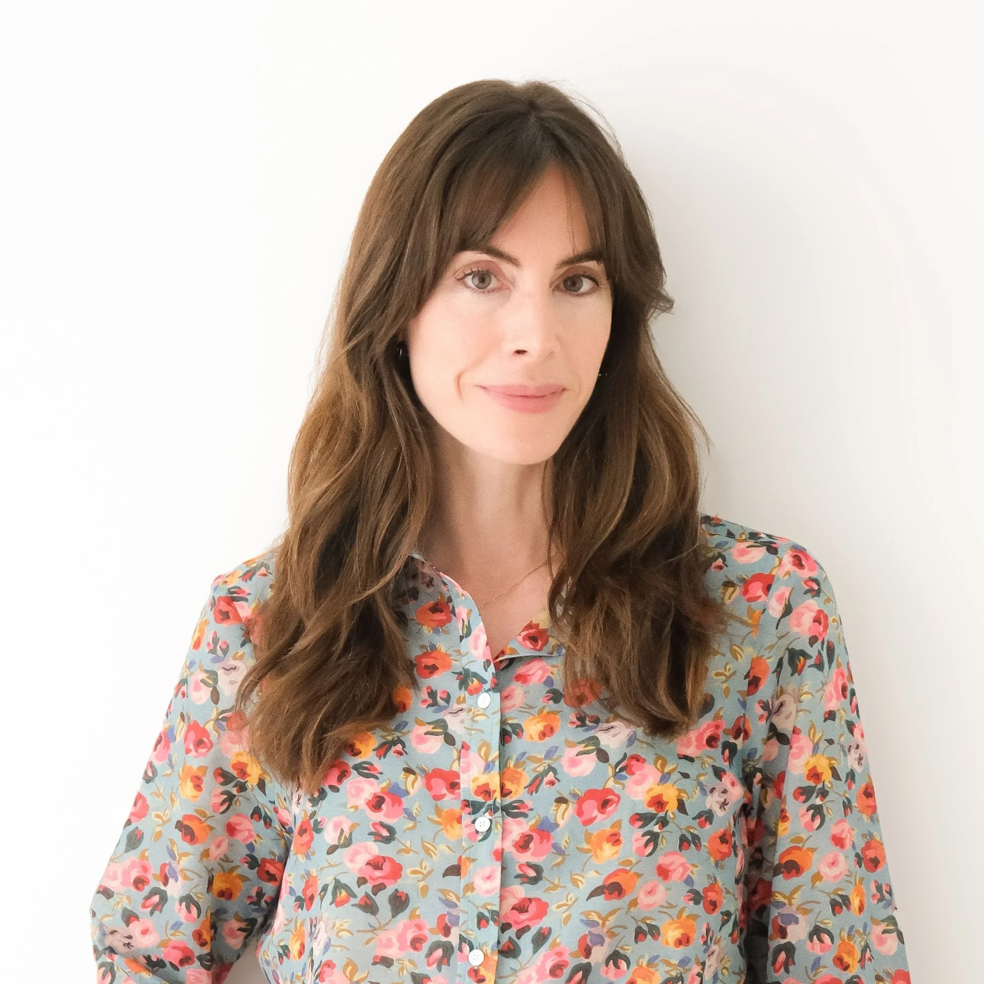 A woman with long brown hair wearing a floral blouse stands against a white background.