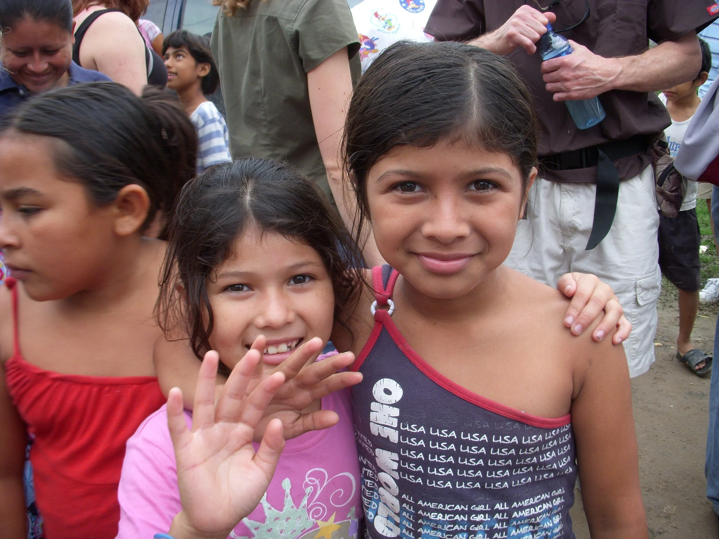 Group of children, two girls in the foreground smiling and waving, at an outdoor event with many people.
