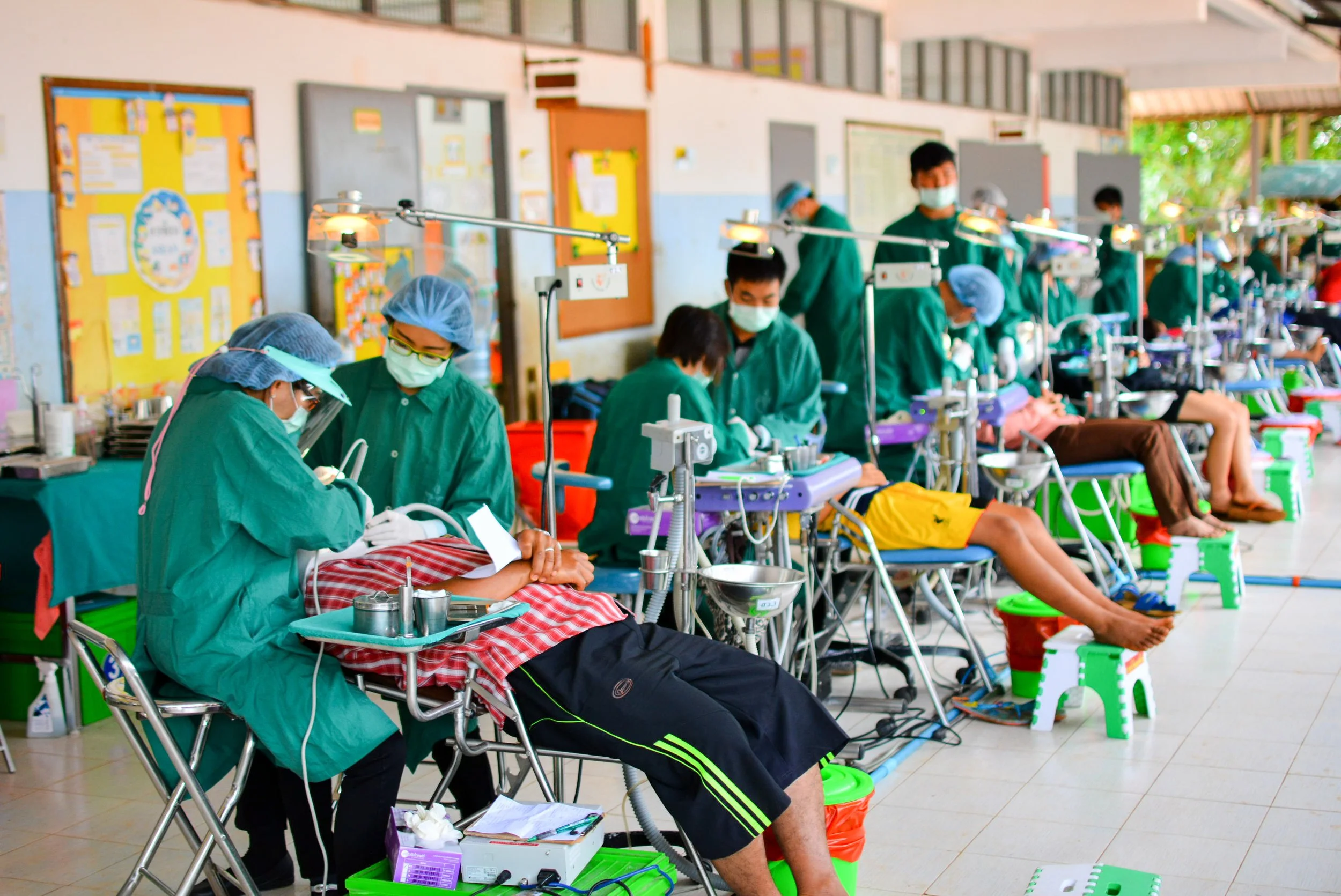 Multiple patients receiving dialysis treatment at a clinic, seated in chairs with medical staff attending to them, inside a well-lit medical facility.