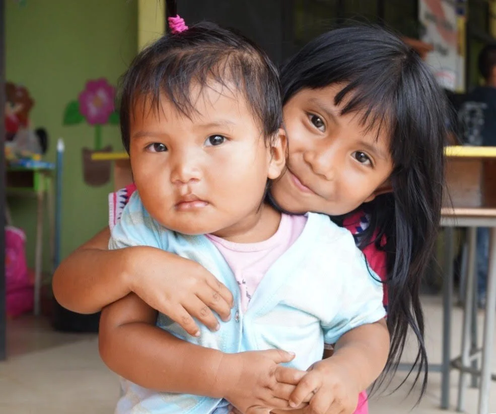 Two young girls, one older and one younger, embracing in a colorful indoor setting with decorated walls and tables.
