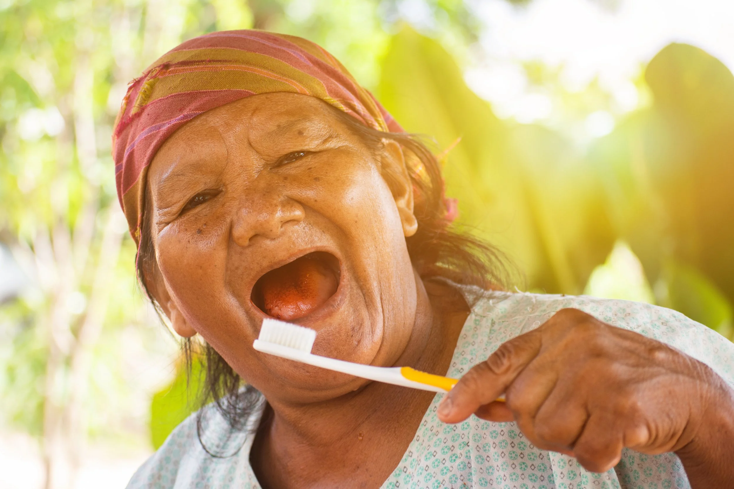 An elderly woman with dark hair, wearing a headscarf and a light patterned top, holding a toothbrush near her open mouth outdoors with green foliage in the background.