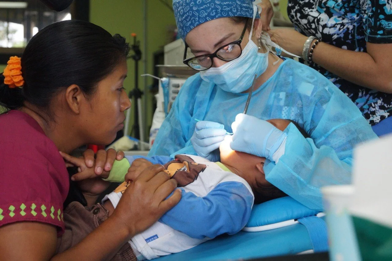 A nurse in scrubs and a mask performs dental surgery on a young child lying on a medical bed, while a woman nearby holds the child's hand and watches intently.