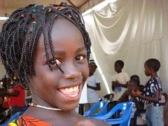 Smiling young girl with braided hair at an indoor event, with other children in the background.