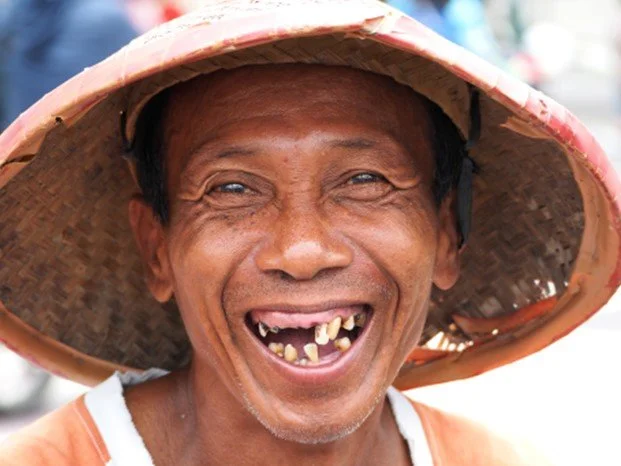 Person wearing a large woven straw hat, smiling widely with missing and crooked teeth, outdoors.