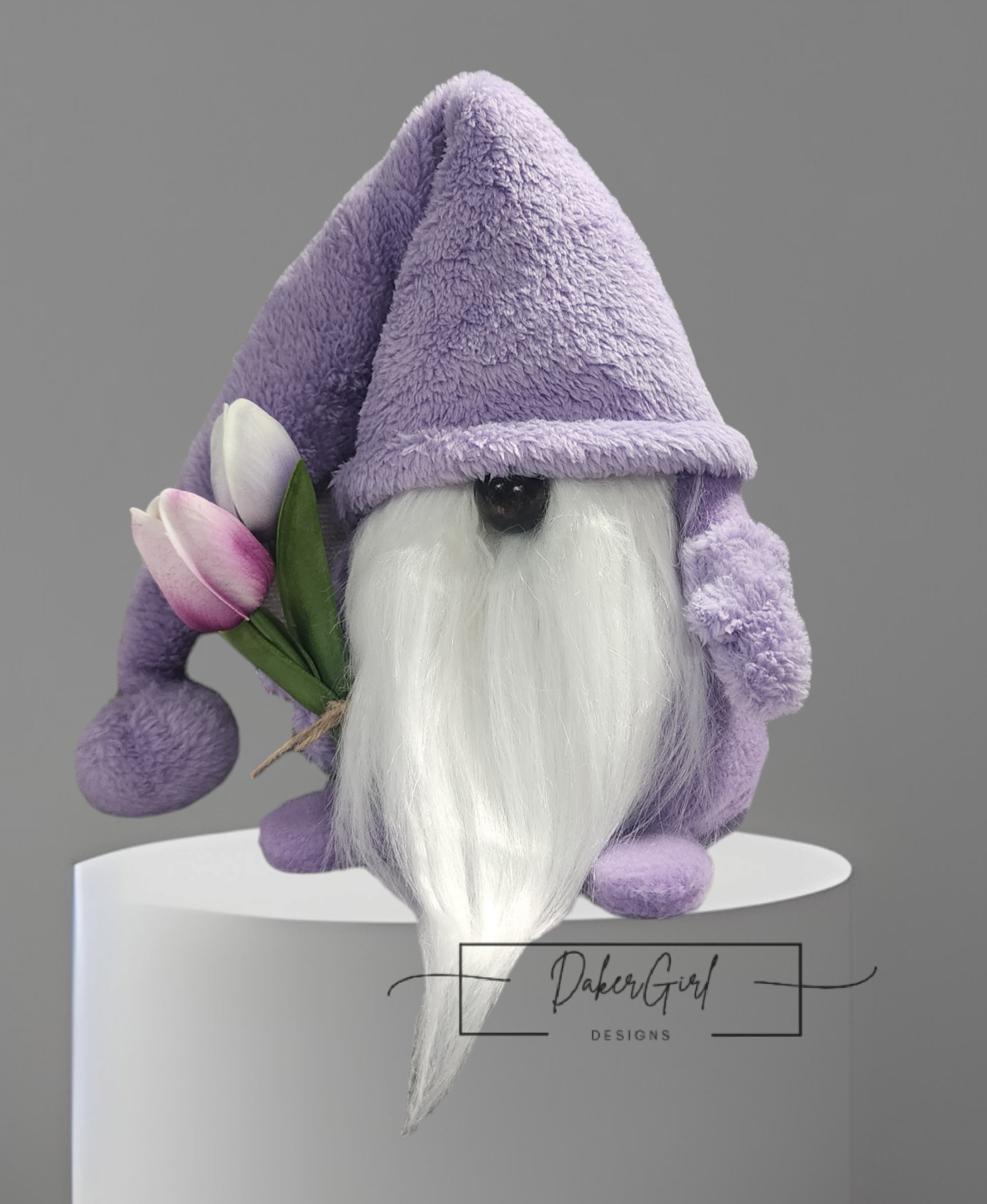 A plush gnome with a long white beard, purple hat and coat, holding pink and white tulips, standing on a white surface with a gray background.
