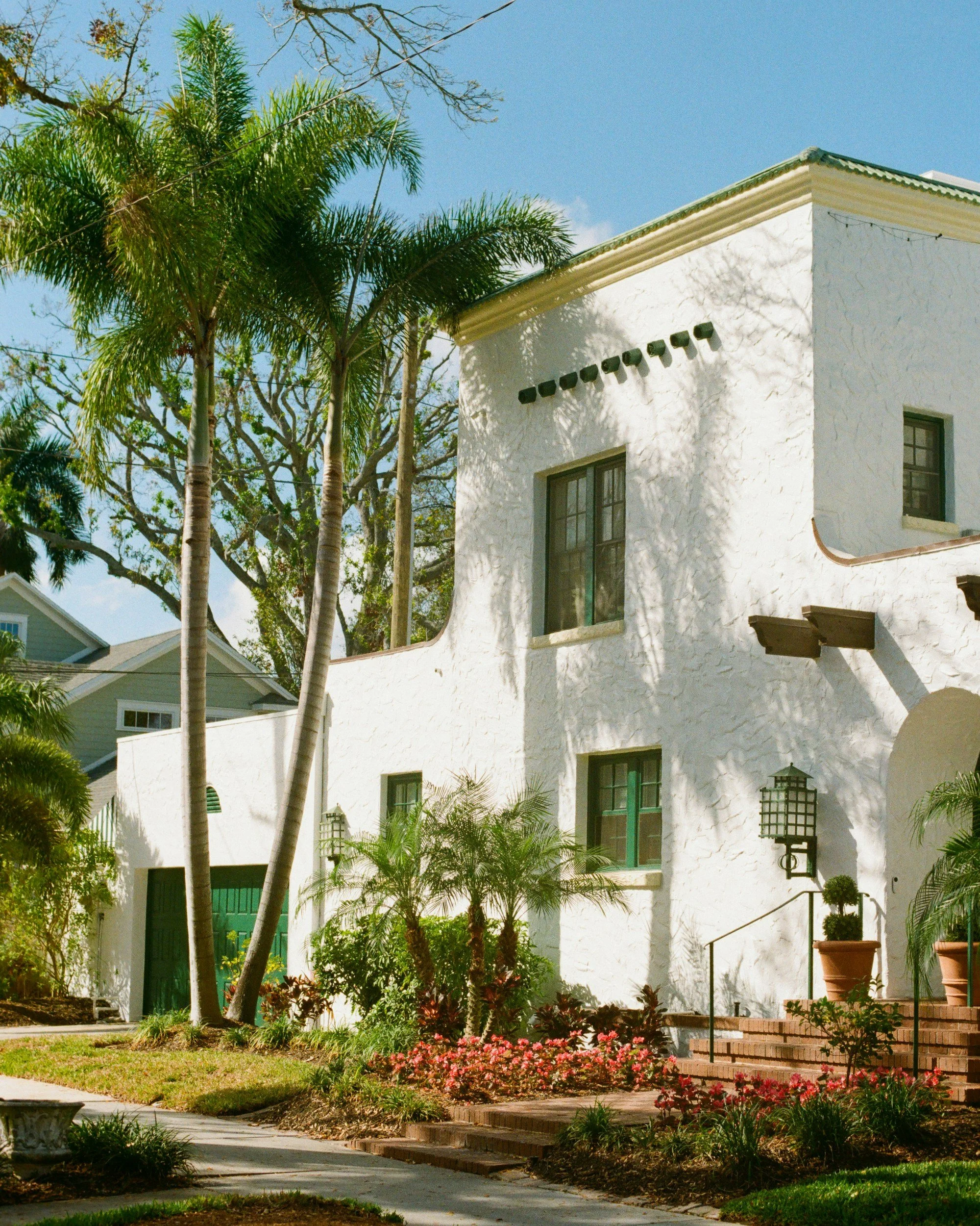 White stucco house with multiple windows, surrounded by lush tropical plants, palm trees, and a garden with flowers, under a clear blue sky.