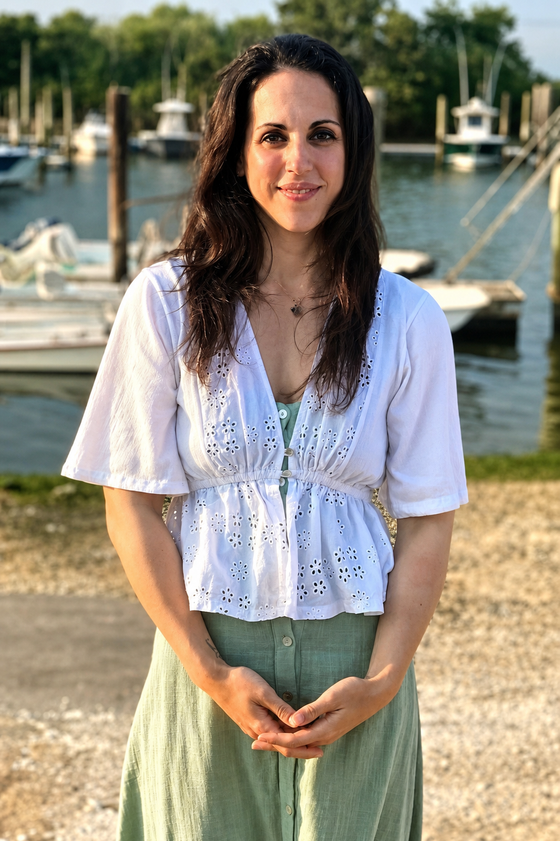 A woman with dark hair standing outdoors near a marina with boats in the background, wearing a white eyelet blouse and a green skirt.