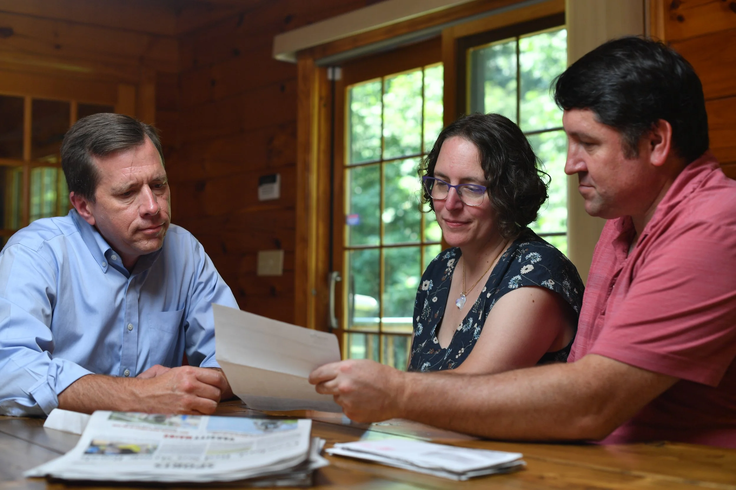 Three adults sitting at a wooden table inside a cabin, reviewing papers with a background of large windows showing greenery outside.