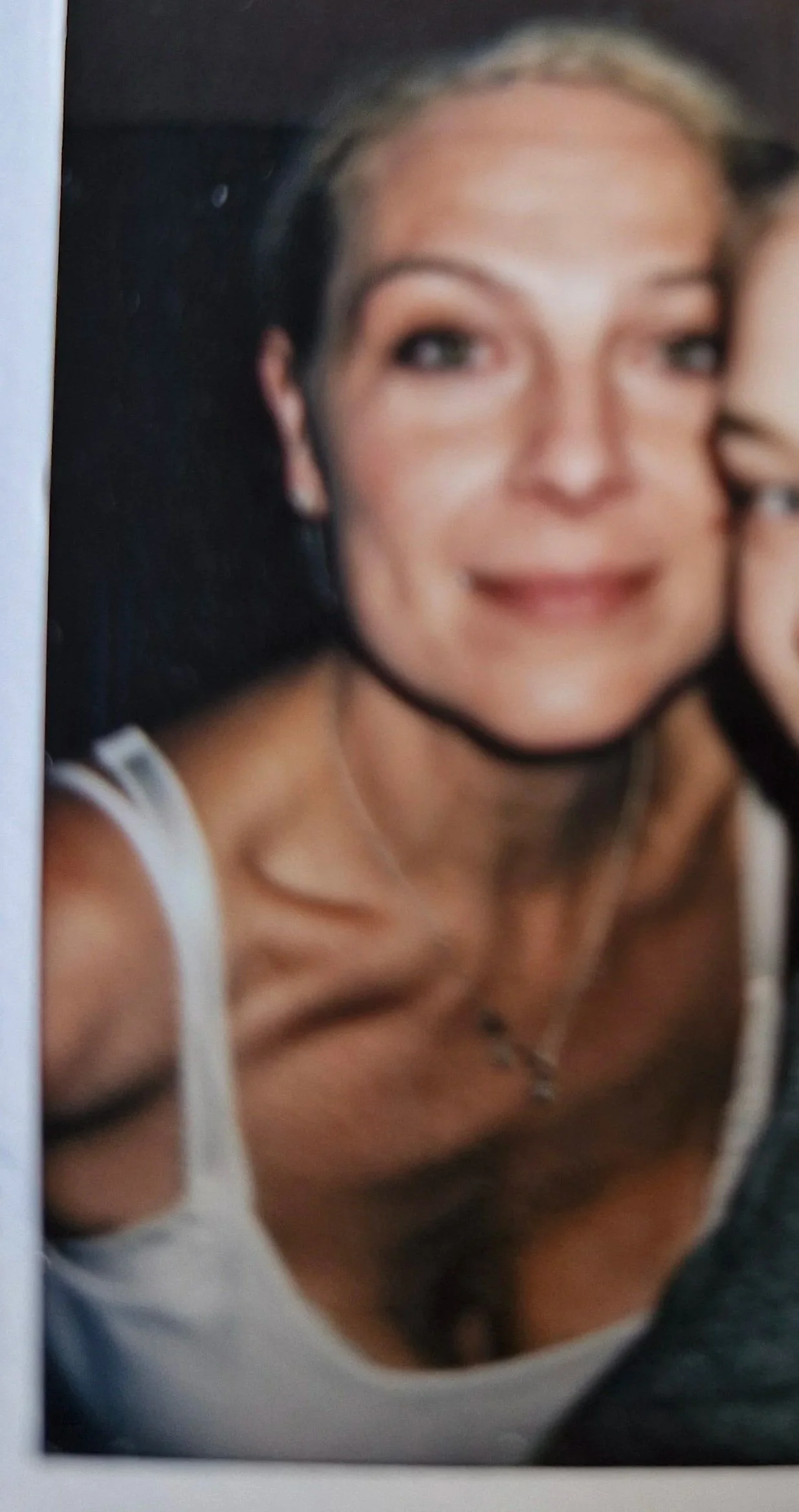 A close-up selfie of a smiling woman with blonde hair and blue eyes, wearing a white tank top, with a blurred background.