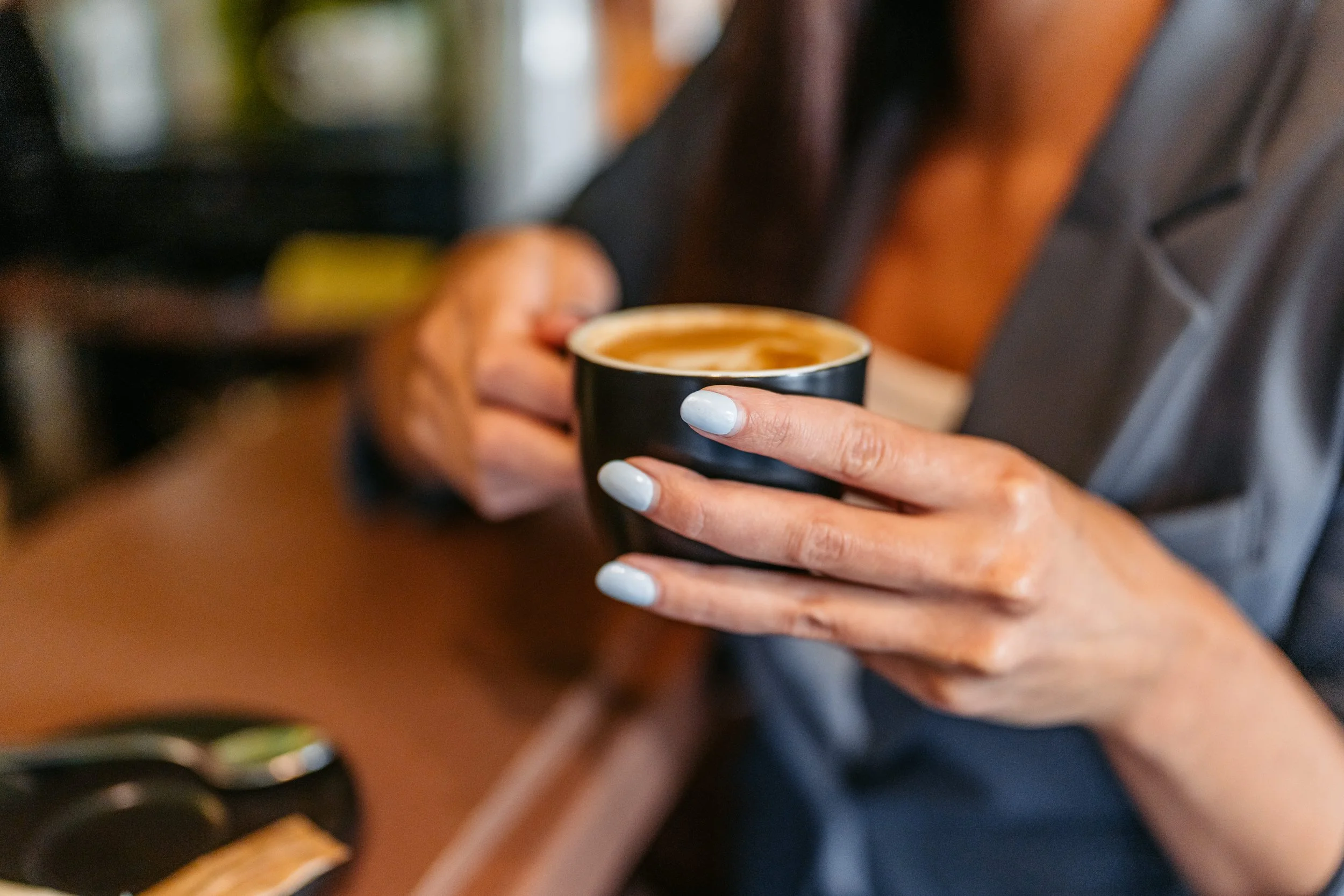 Person holding a black coffee mug with latte art, sitting at a wooden table.