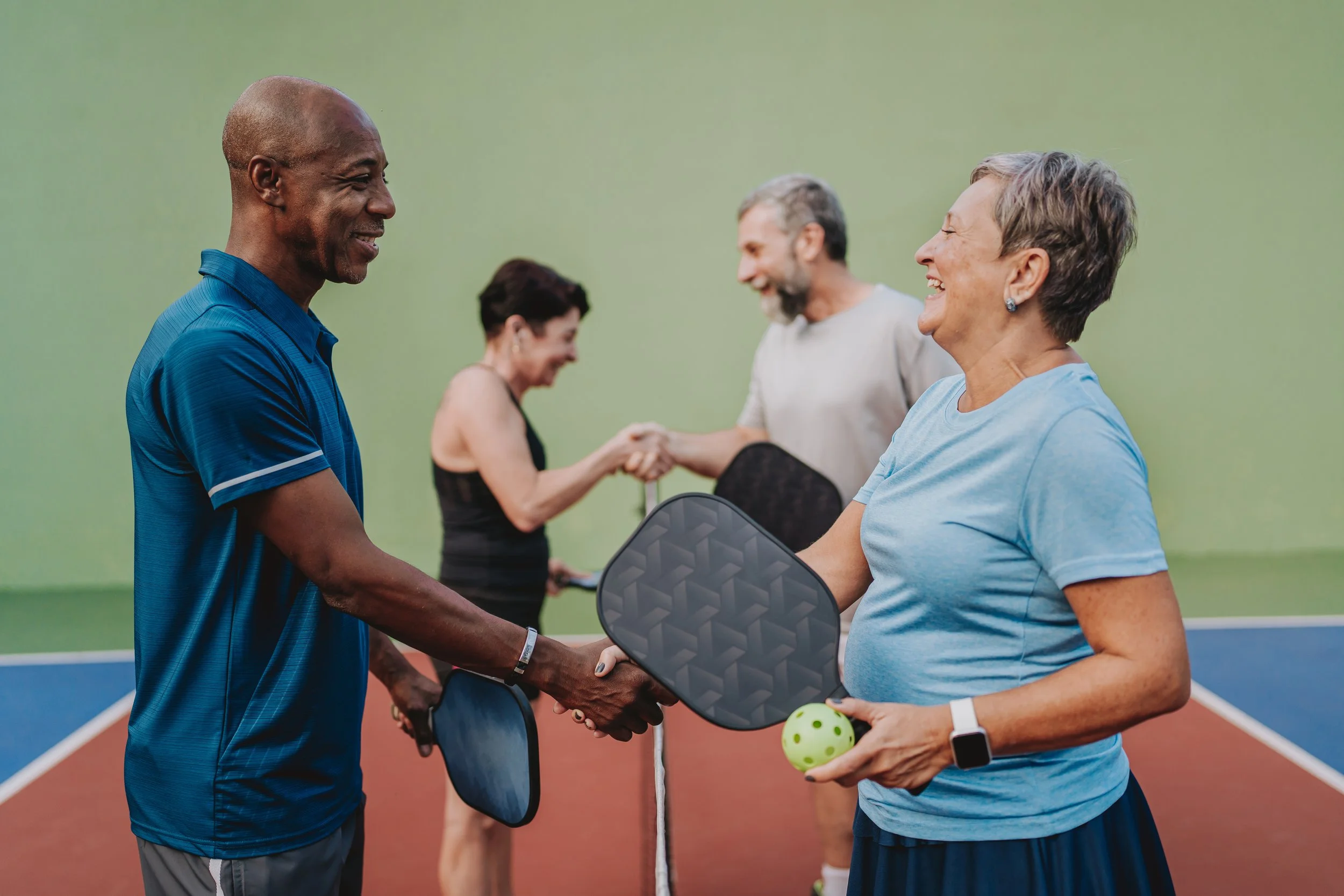 A group of older adults on a tennis court, shaking hands and smiling, with two holding pickleball paddles and ball.