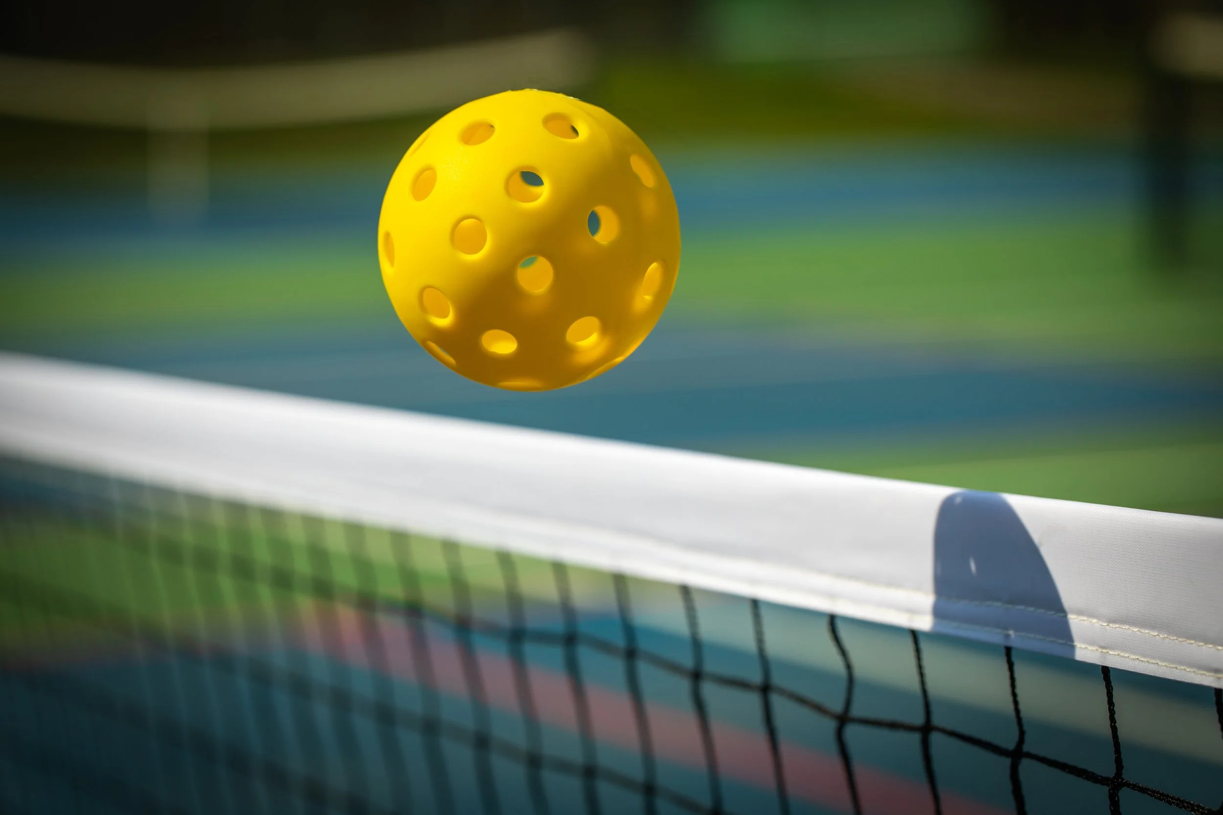 Yellow perforated tennis ball in mid-air near a tennis net on a tennis court.
