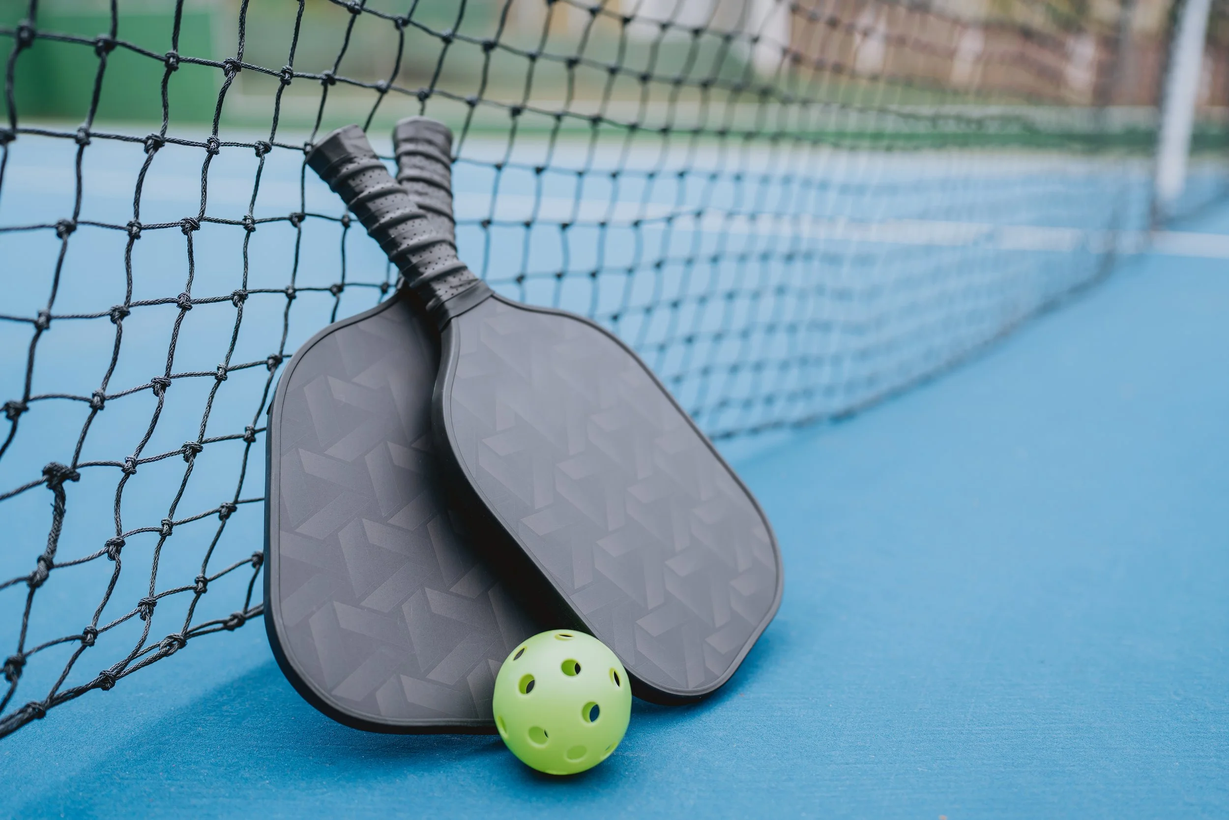 Two paddle rackets, one yellow perforated ball, on a blue outdoor tennis or pickleball court near a black net.