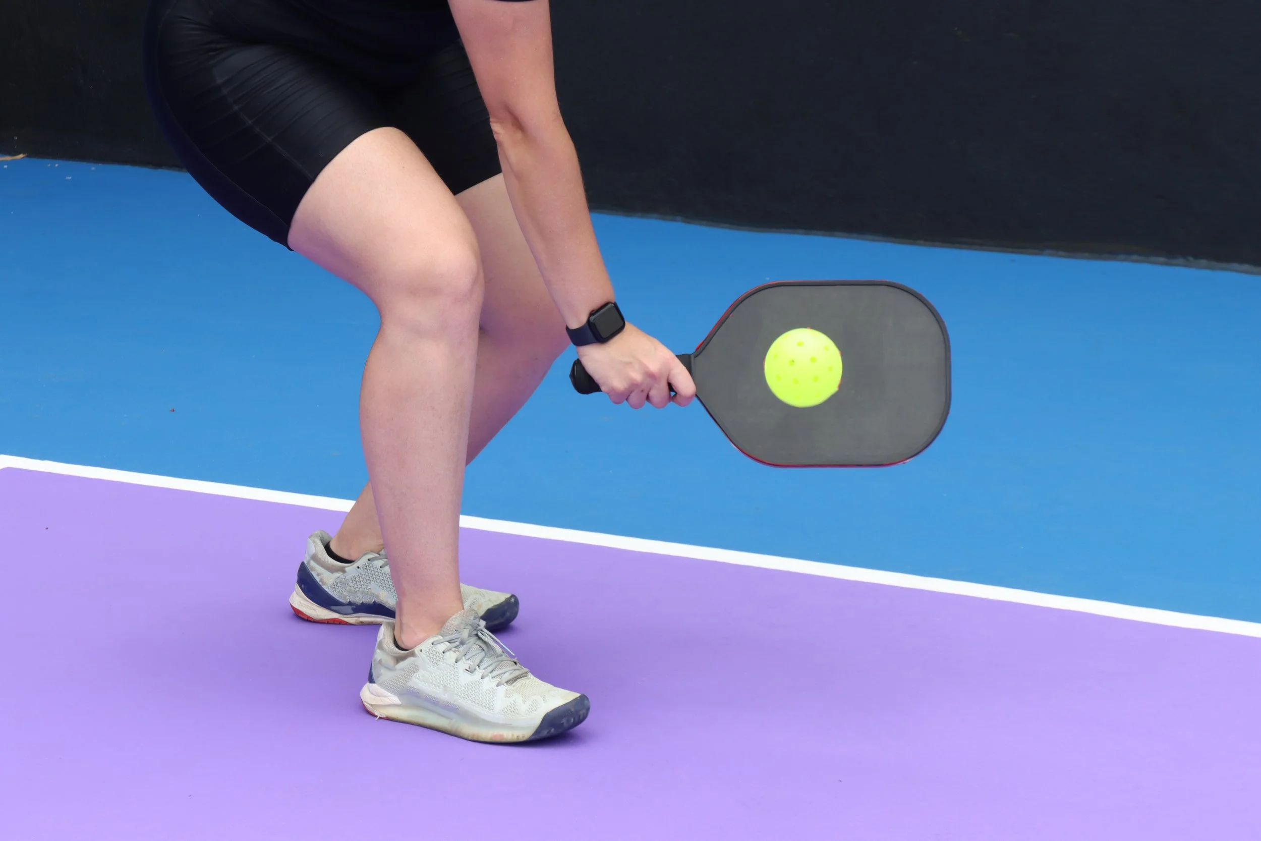Person in athletic clothing preparing to hit a yellow ball with a pickleball paddle on a purple and blue court.