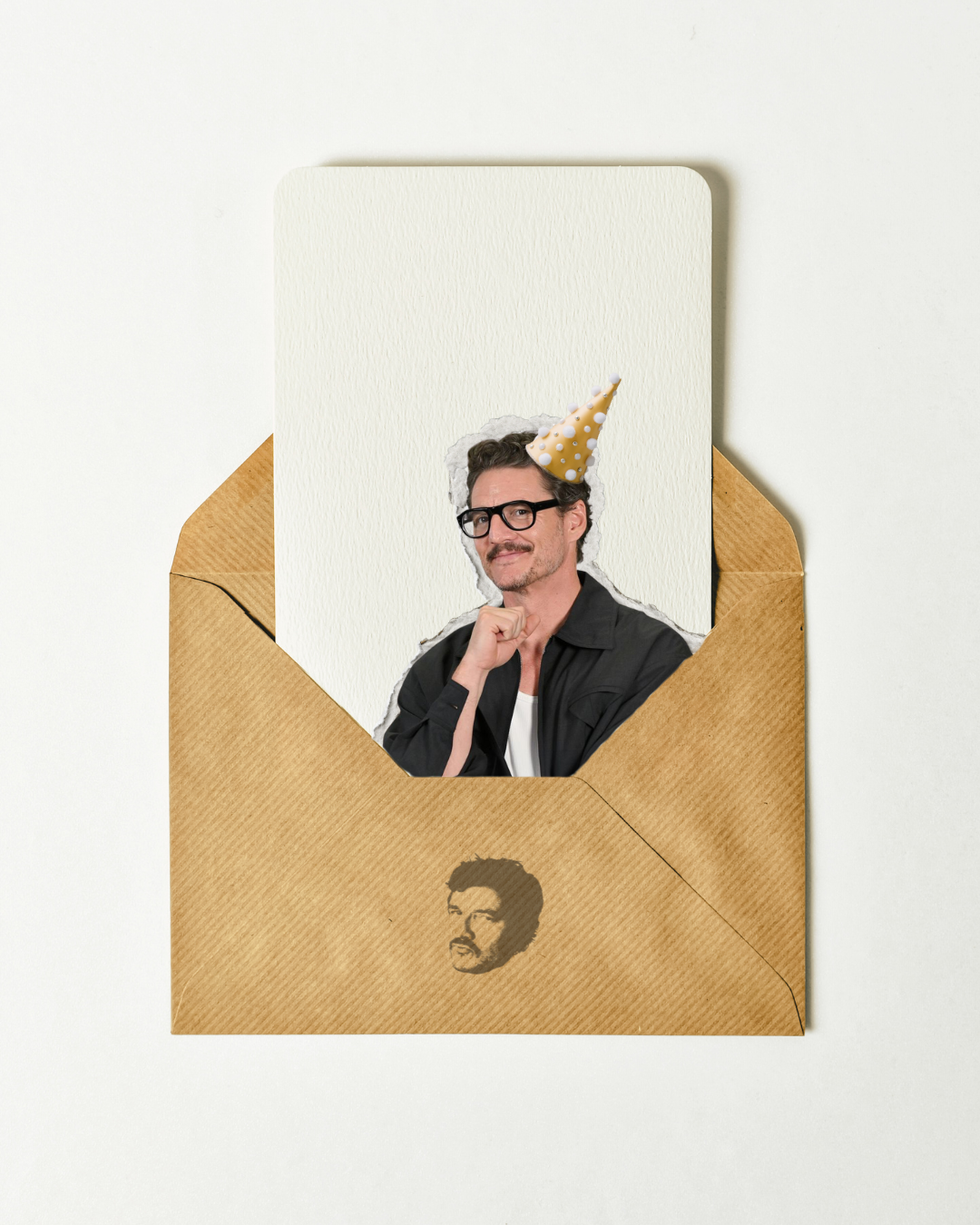 A man wearing glasses and a birthday hat, sitting with his chin resting on his hand, inside a paper envelope with a printed image of a face on it.