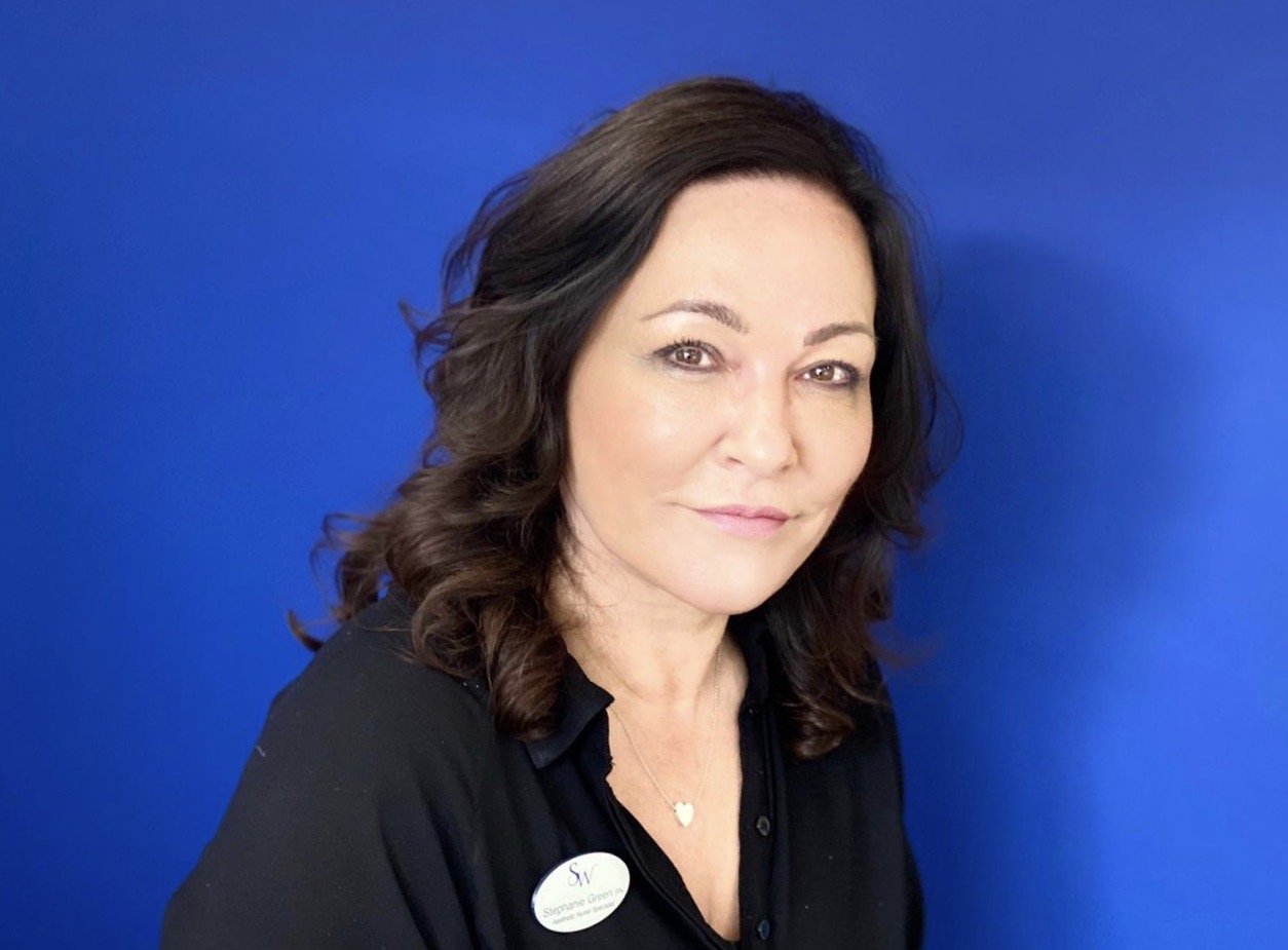 Portrait of a woman with dark, wavy hair and wearing a black top, standing in front of a blue background.