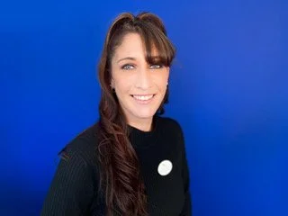 A woman with long brown hair, wearing a black top, smiling in front of a blue background.