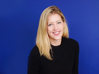 Portrait of a woman with blonde hair wearing a black top, smiling against a blue background.