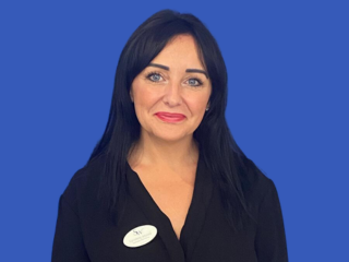 Portrait of a woman with long black hair wearing a black top and a name badge, standing against a blue background.