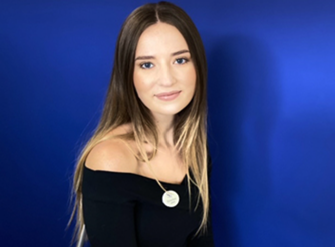 A young woman with long, light brown hair and fair skin, wearing an off-the-shoulder black top, standing against a solid blue background.