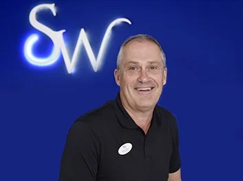 A smiling man with short gray hair wearing a black polo shirt, standing in front of a blue background with illuminated white letters 'SW'.