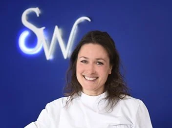 A woman with shoulder-length dark hair smiling, wearing a white top, standing in front of a blue wall with a lit logo displaying the letters S and W.