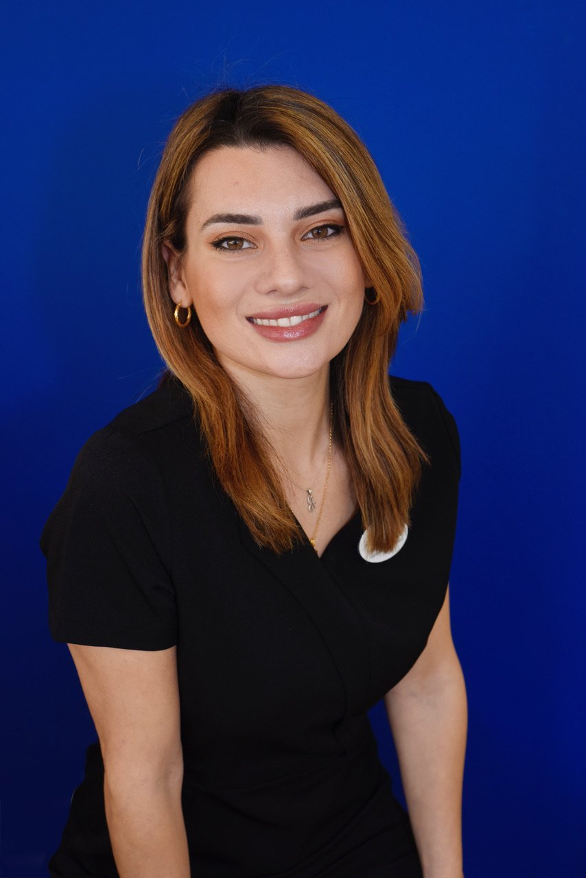 Portrait of a smiling woman with light brown hair, wearing a black top, gold earrings, and a gold necklace, against a solid blue background.