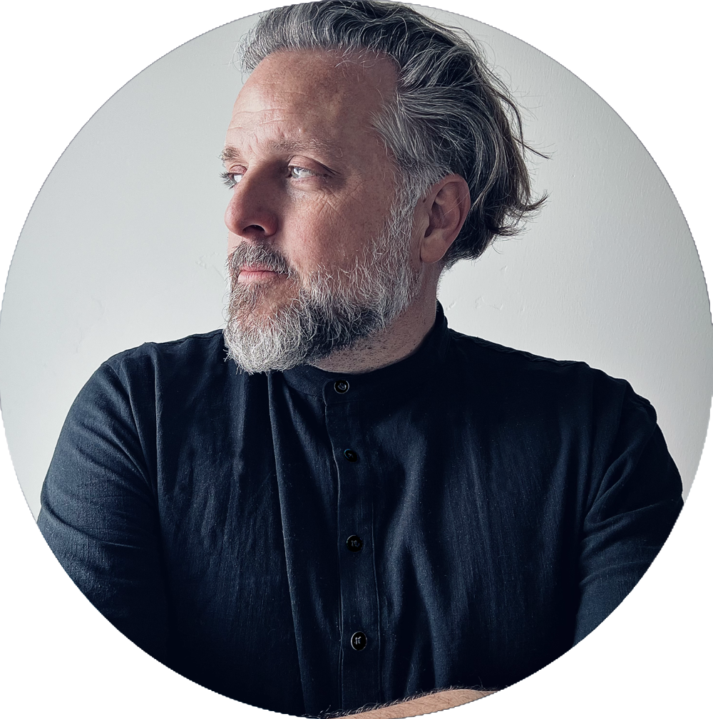 A middle-aged man with gray hair and beard, wearing a black button-up shirt, looking to his left against a plain white background.
