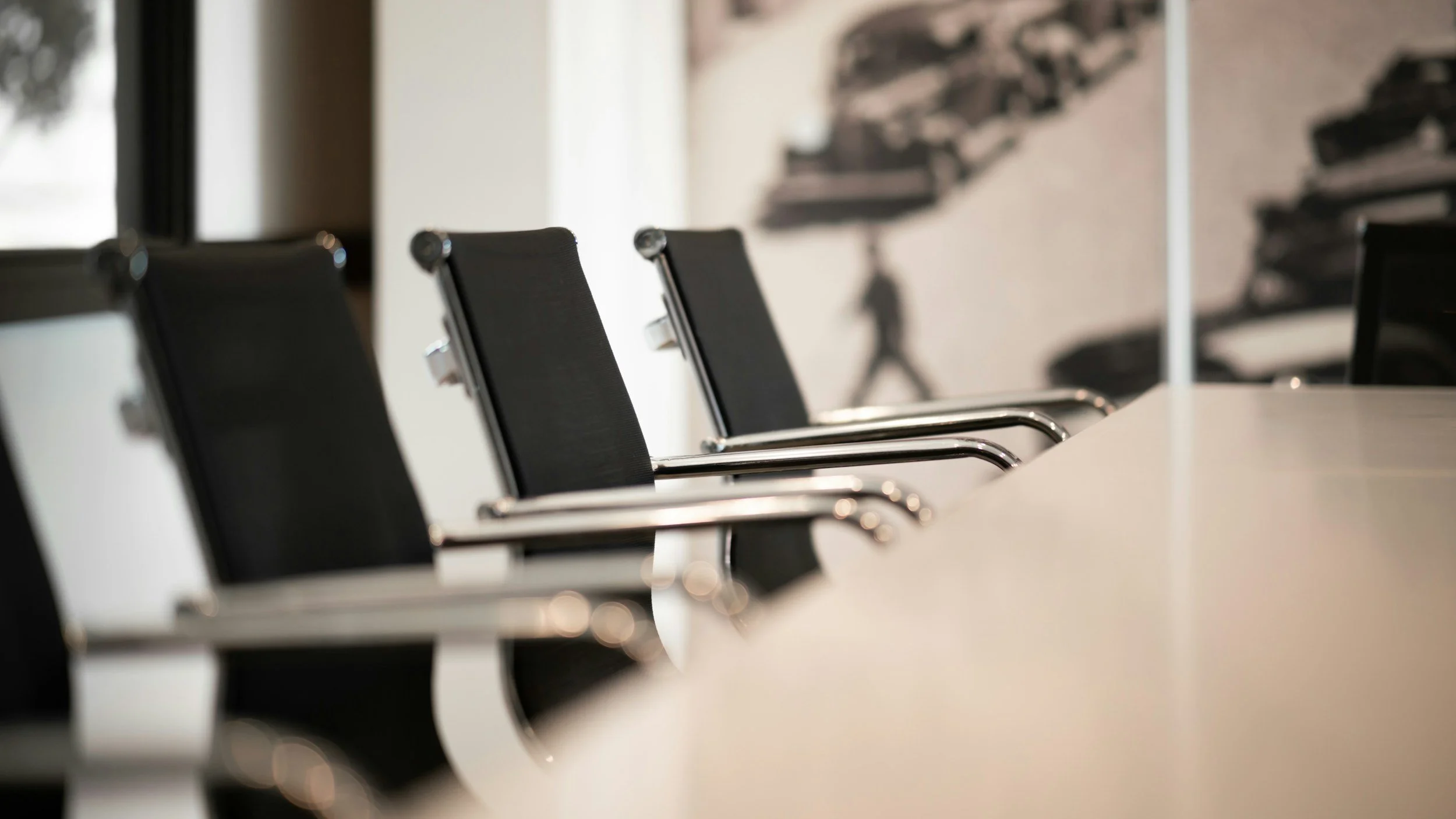 Empty black conference room chairs with chrome armrests lined up at a long white table in a modern office, with a blurry wall art of stacks of books and a person walking in the background.